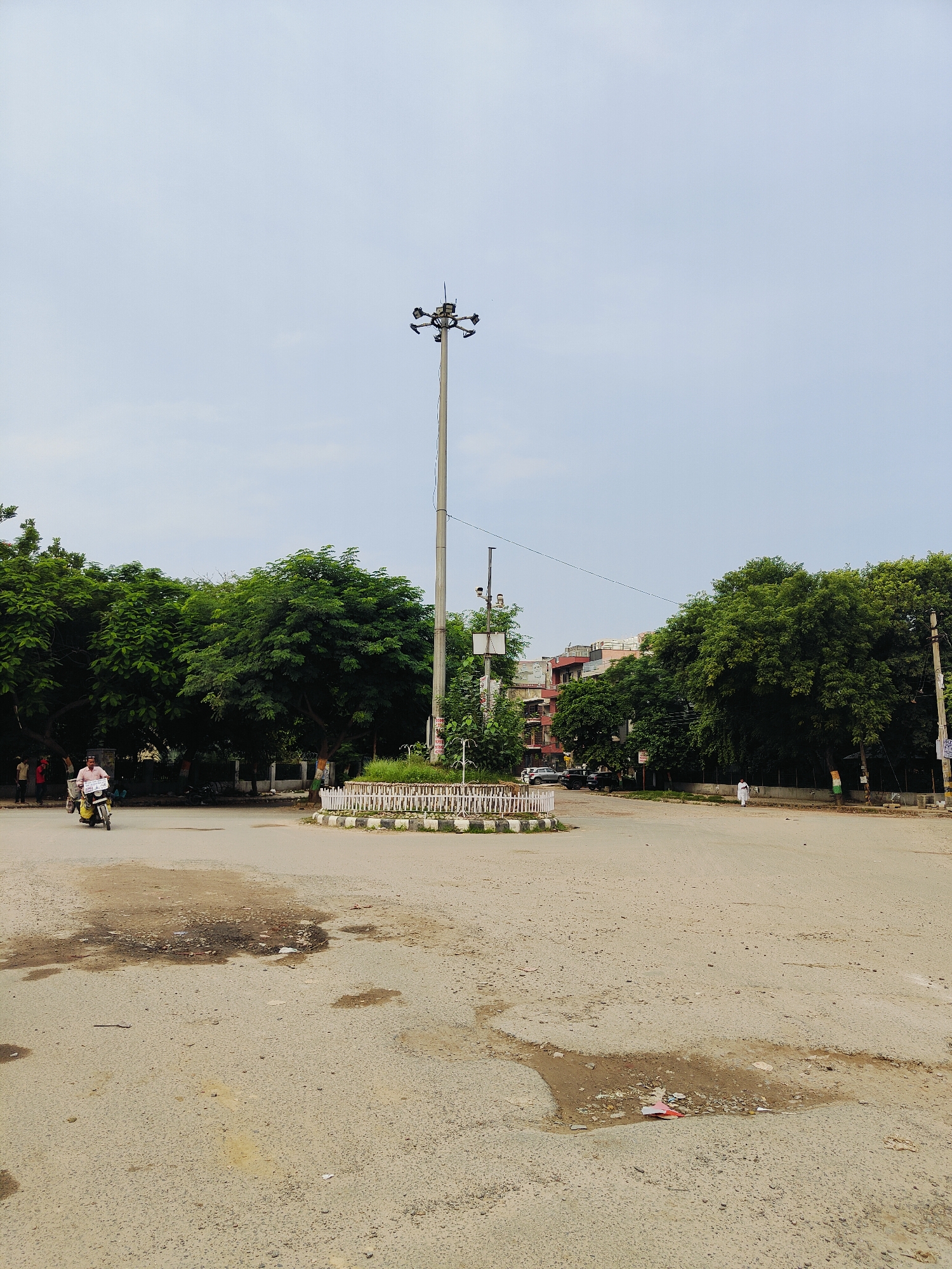 In Gurgaon, India, a wide, paved intersection features a prominent tall lamppost with multiple lights at its apex, positioned near a circular, white-railed planter filled with green vegetation. To the left, a man on a yellow scooter, holding a white object, rides on the asphalt surface, which shows some signs of wear and water puddles. Behind him, a cluster of lush green trees forms a dense backdrop, with several people visible in the shade of the trees. Further back, a multi-story building with red-tiled roofs is situated amongst more trees and other structures. To the right, the road continues, lined by trees and buildings, with another lamppost and various signs visible. The sky above is overcast and light blue.