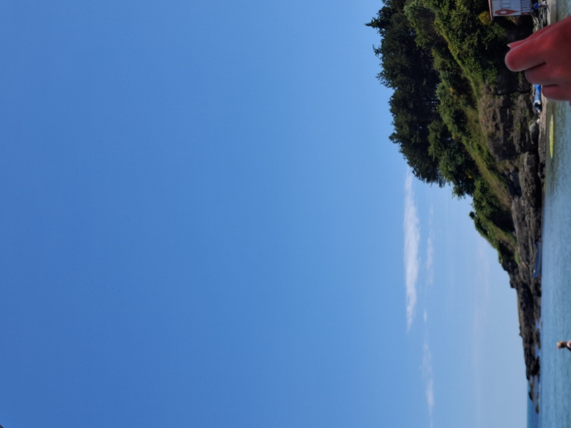 A clear blue sky dominates the upper portion of the image. Below, a rocky shoreline is visible, covered in lush green vegetation and trees. The sea is a calm blue. In the foreground, a blurred hand is reaching into the frame from the right. A small white and red structure is partially visible on the right edge of the shore. A person is standing in the water near the shore, and a small blue boat is moored near the rocky outcrop. The scene appears to be a coastal area on a sunny day.
