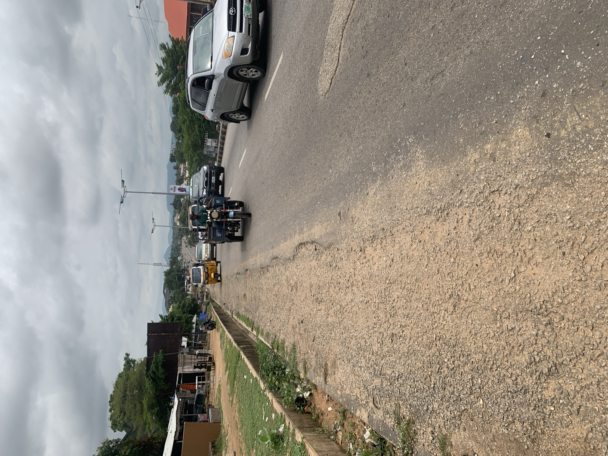 The image depicts a cloudy day scene on a street in Adankolo, Nigeria. In the foreground, a rough, gravelly roadside runs along the left side of the frame, separated from the paved road by a concrete drainage channel that contains some debris. On the paved road, several vehicles are visible, moving in the same direction. Closest to the camera on the right is a silver SUV, followed by a dark SUV. Further ahead, a yellow auto-rickshaw (Keke Napep) is in motion, with another auto-rickshaw behind it. The road itself shows signs of wear and tear, with visible potholes and uneven patches. On the left side of the road, there are several modest structures, appearing to be market stalls or small shops, with corrugated metal roofs and open fronts. Lush green trees and vegetation are present behind these structures, and in the background, hills and mountains are visible under the overcast sky. The overall impression is that of a busy but somewhat unkempt urban environment.