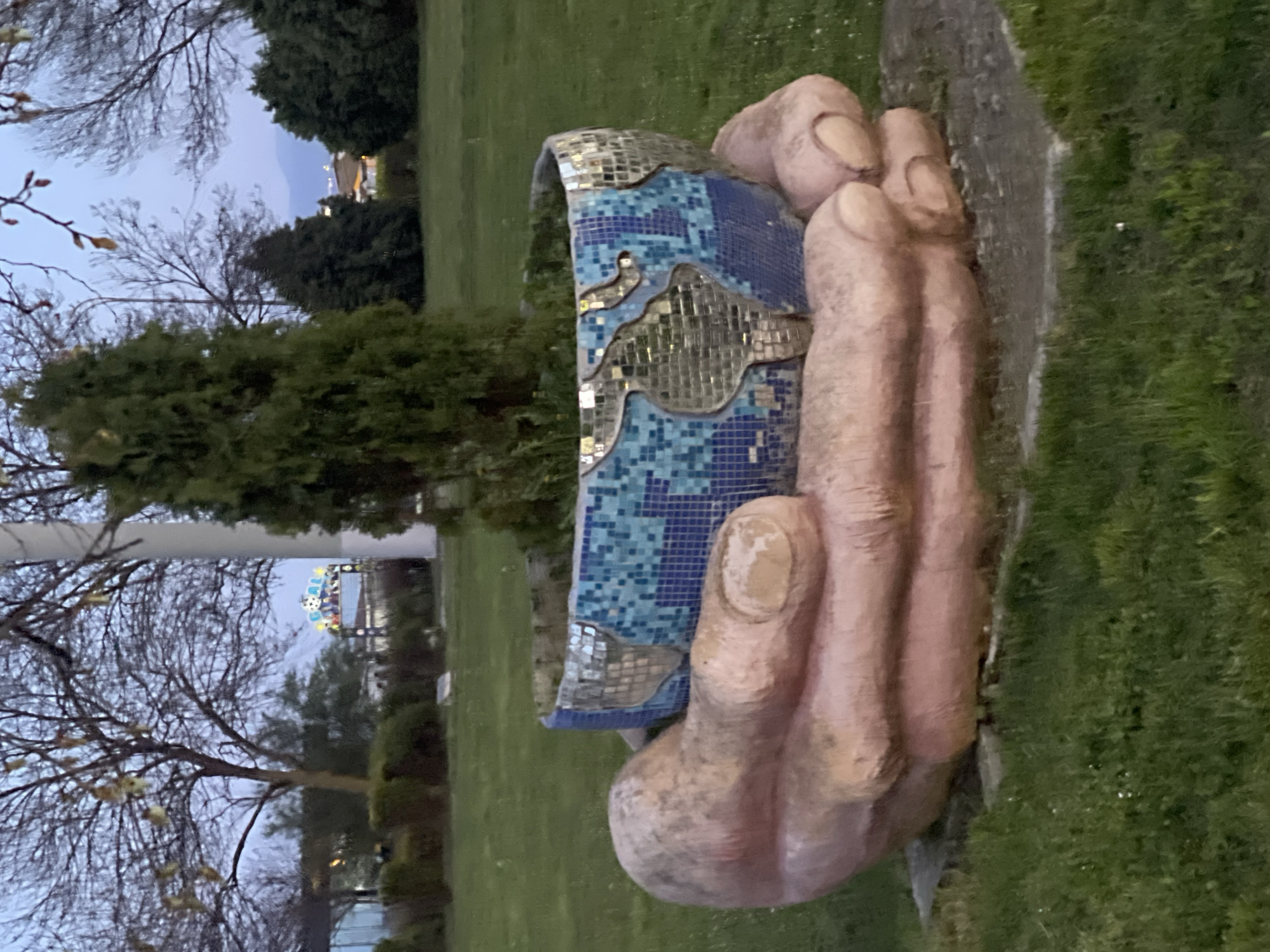 A vertical, medium shot depicts a large sculpture of a hand holding a blue and silver mosaic object. The hand is peach-colored and appears to be made of stone or concrete. The mosaic object has a curved shape and features a pattern of blue and silver tiles, resembling a map of the world. The sculpture is situated on a grassy lawn. In the background, trees and a building with lights are visible. The sculpture is in Burgas, Bulgaria.