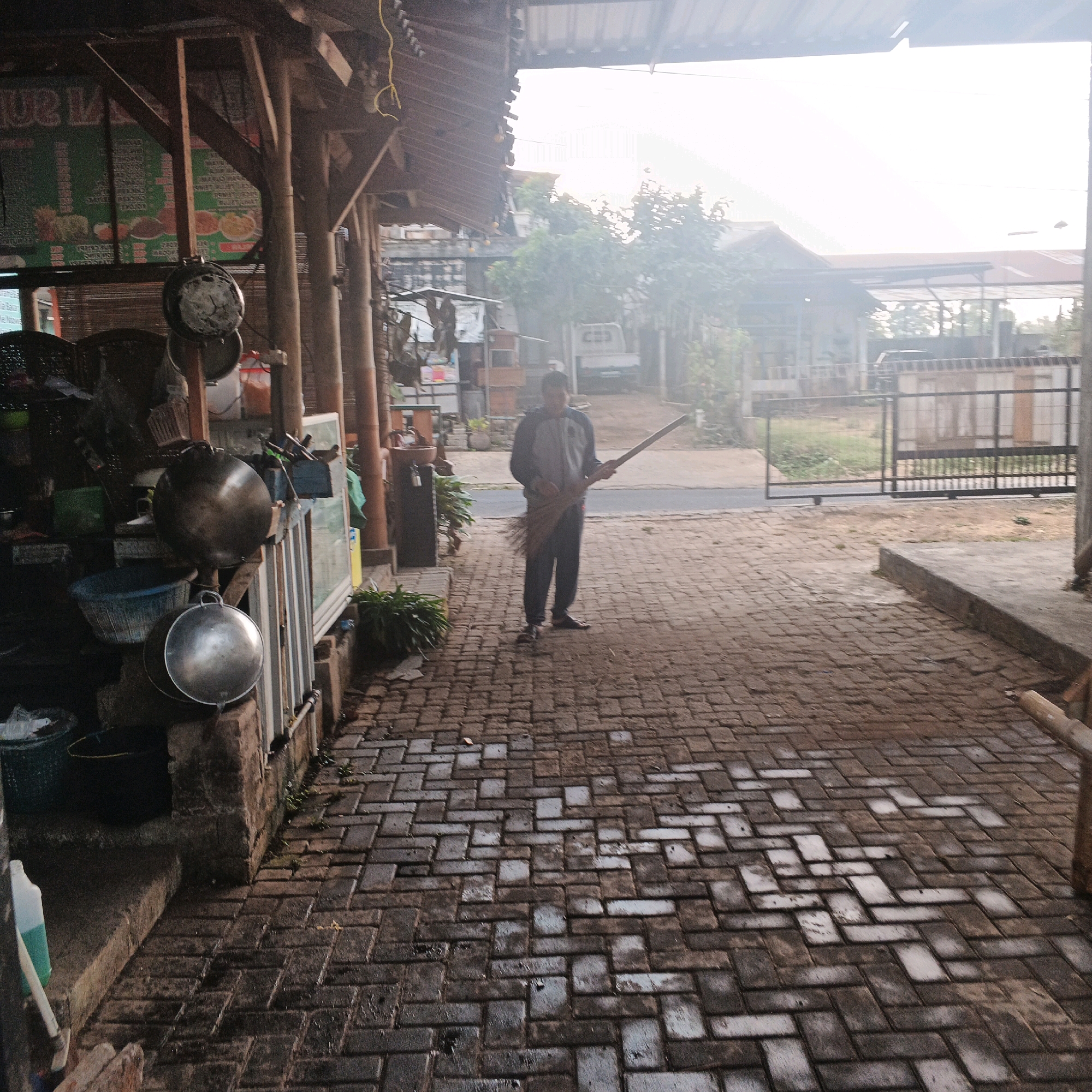 The image depicts an outdoor scene in what appears to be a commercial or residential area, possibly a marketplace or a roadside stall, in Bocek, Indonesia. A person is standing in the middle of a brick-paved pathway, holding a broom and facing away from the viewer. The person is wearing a grey long-sleeved shirt and dark trousers. To the left of the pathway, there is a wooden structure with a roof, supported by poles. Underneath this structure, various items are visible, including hanging pots and pans, baskets, and containers, suggesting a food stall or a shop selling kitchenware. The pathway is wet, indicating it may have recently rained or been cleaned. In the background, there are buildings, trees, and a metal gate. The overall lighting suggests it is daytime, possibly early morning or late afternoon, with some atmospheric haze.