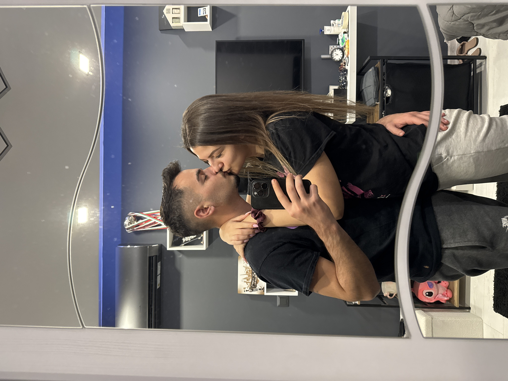 A couple is kissing and taking a selfie in front of a mirror inside an apartment. The man is wearing a black t-shirt and gray sweatpants. The woman is wearing a black t-shirt and gray sweatpants. Behind them, there is a TV, shelves with various objects, and a gray wall with decorations. There is also a humidifier visible. The location cannot be determined as Sofia, Bulgaria, based on the image.