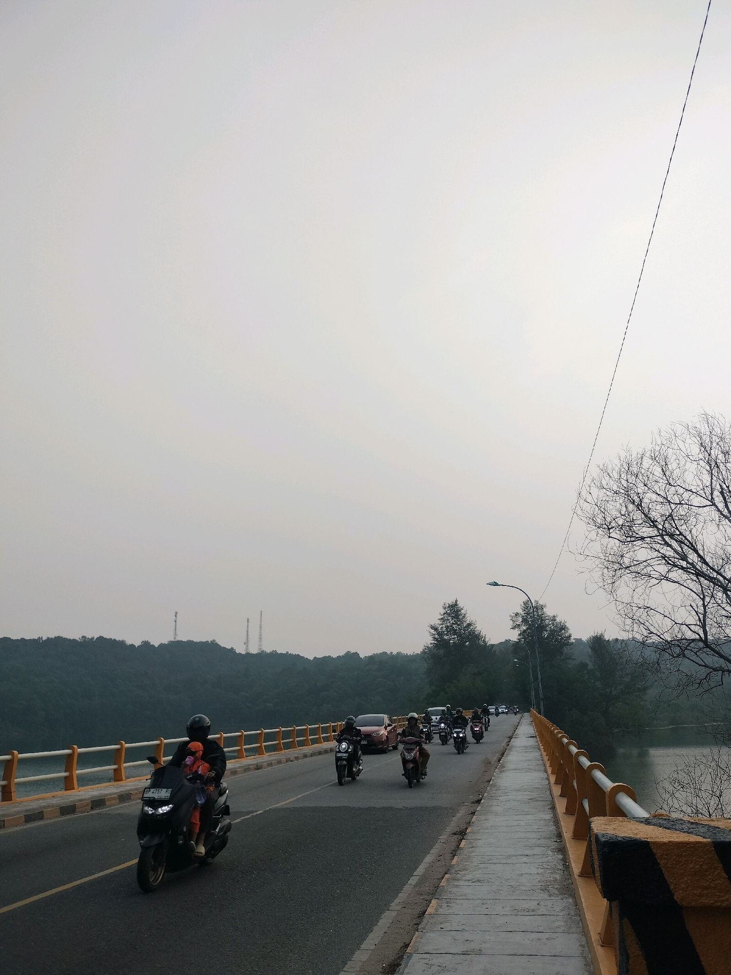 The image depicts a bridge scene, likely in Batam, Indonesia, during overcast daylight. A line of motorcycles and a few cars are traveling across the bridge. On the left side of the bridge, a black motorcycle with a rider wearing a helmet and orange clothing is prominent in the foreground. The rider is carrying a child also dressed in orange. Further down the road, more motorcycles are visible, some with multiple riders. On the right side of the bridge, there is a pedestrian walkway with a yellow and black striped railing. Beyond the railing, a body of water, possibly a lake or reservoir, is visible, bordered by a dense green forest and hills in the background. Two tall tower-like structures are visible on the distant hills. A single bare tree branch is in the upper right corner of the frame, and a streetlight is positioned over the road. The sky is a uniform light gray, suggesting a hazy or cloudy atmosphere.