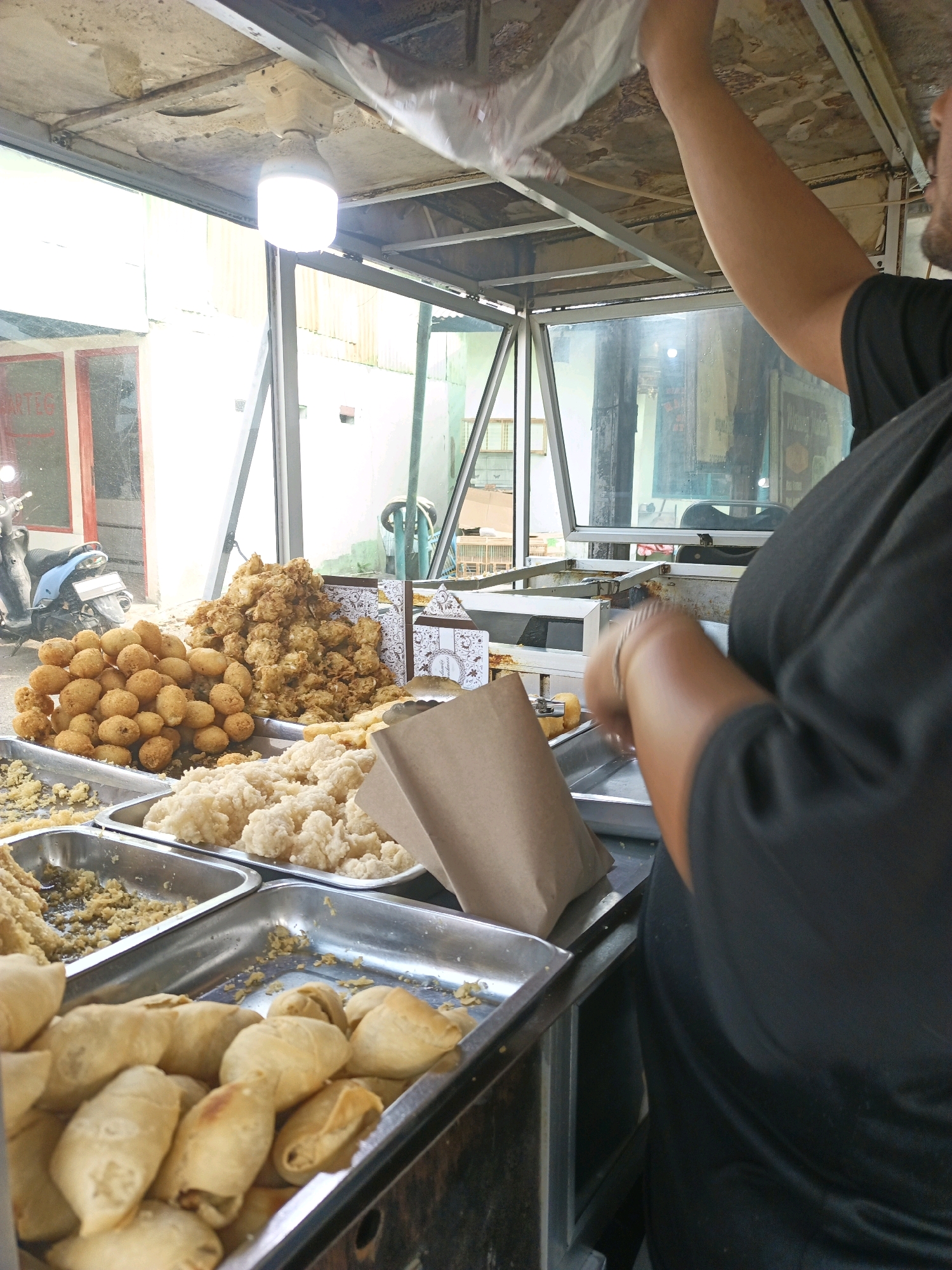 An overhead shot captures a street food stall in South Jakarta, Indonesia, with a vendor in the foreground preparing food. The stall, a covered structure with metal framing and a lightbulb overhead, displays a variety of fried snacks. Trays overflow with golden-brown fried items, including what appear to be small, round fritters, larger, more irregularly shaped fried pieces, and lighter-colored, fluffy fried snacks. In the foreground, a tray holds several fried spring roll-like items. A brown paper bag sits on one of the trays. The vendor, wearing a black t-shirt, is partially visible on the right, with their arm raised, holding a plastic bag over the food. In the background, through the glass front of the stall, a blue motorcycle is parked outside. The scene suggests a bustling outdoor market or roadside eatery.