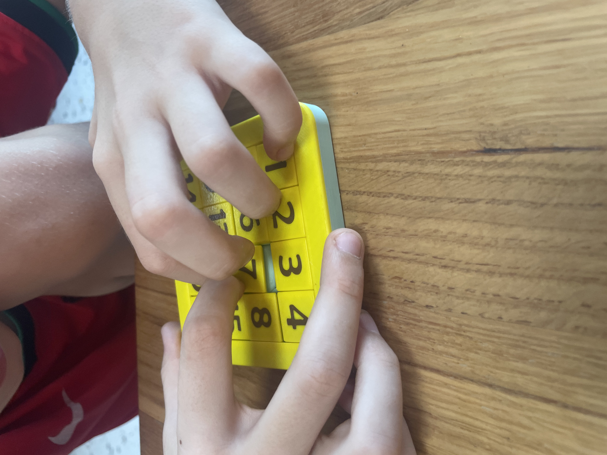 In this close-up shot, two children's hands are engaged with a yellow sliding puzzle game on a wooden table. The puzzle has numbered tiles, with visible numbers including 1, 2, 3, 4, 5, 7, and 8. One child's left hand is positioned to the lower left of the puzzle, while their right hand, along with another child's hands, manipulates the tiles. The children are wearing red shirts. The background is blurred, but hints at a domestic setting, potentially a home in Bac Nha Trang Ward, Vietnam.