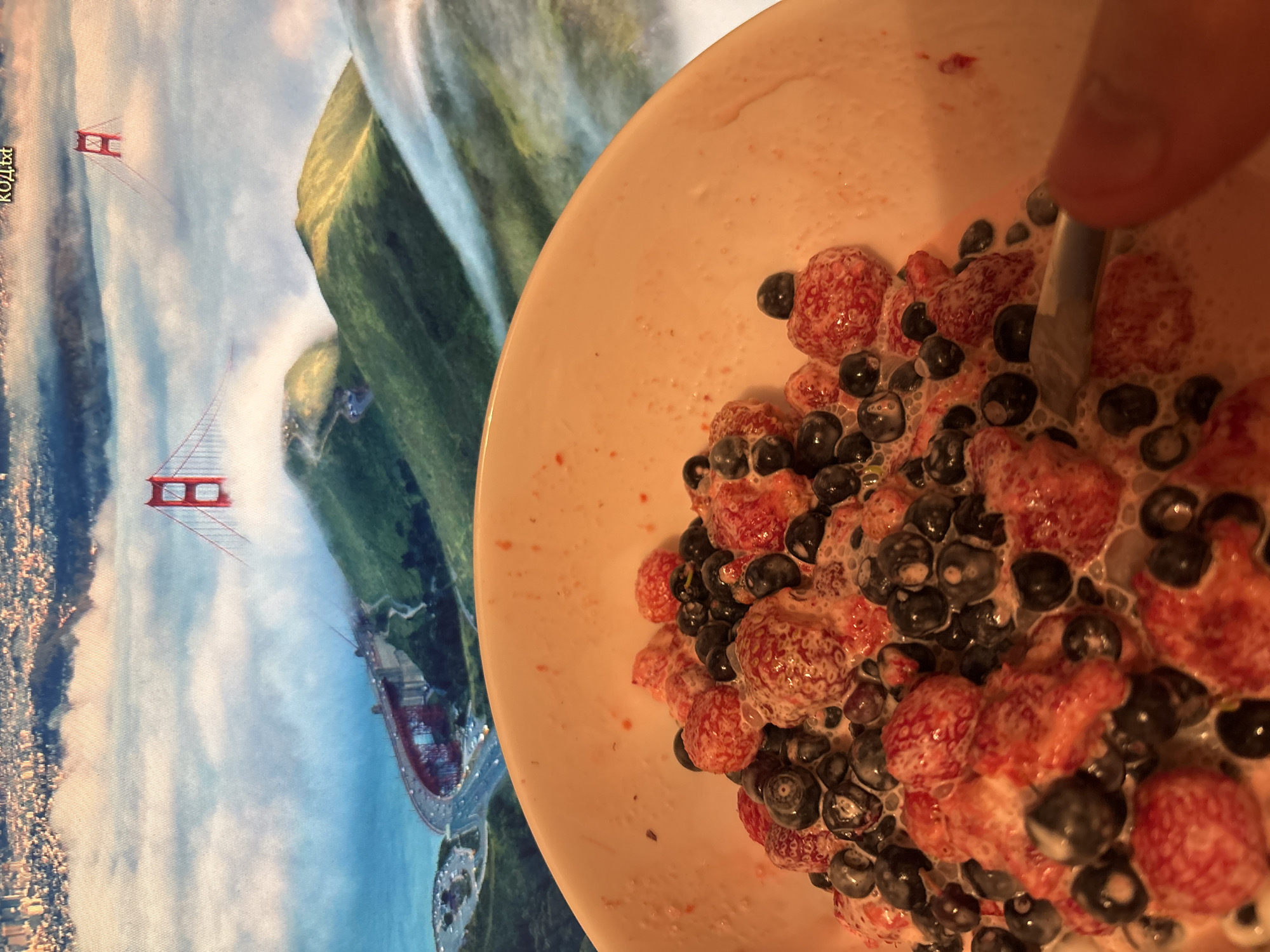 A close-up shot focuses on a bowl filled with strawberries and blueberries in a creamy liquid. In the background, a scenic view of the Golden Gate Bridge in San Francisco is displayed, with the iconic red towers emerging from a bed of clouds that partially obscure the green hillsides and the city skyline. A person's thumb and a metallic utensil are visible at the bottom right of the frame, suggesting that the food is being prepared or consumed.