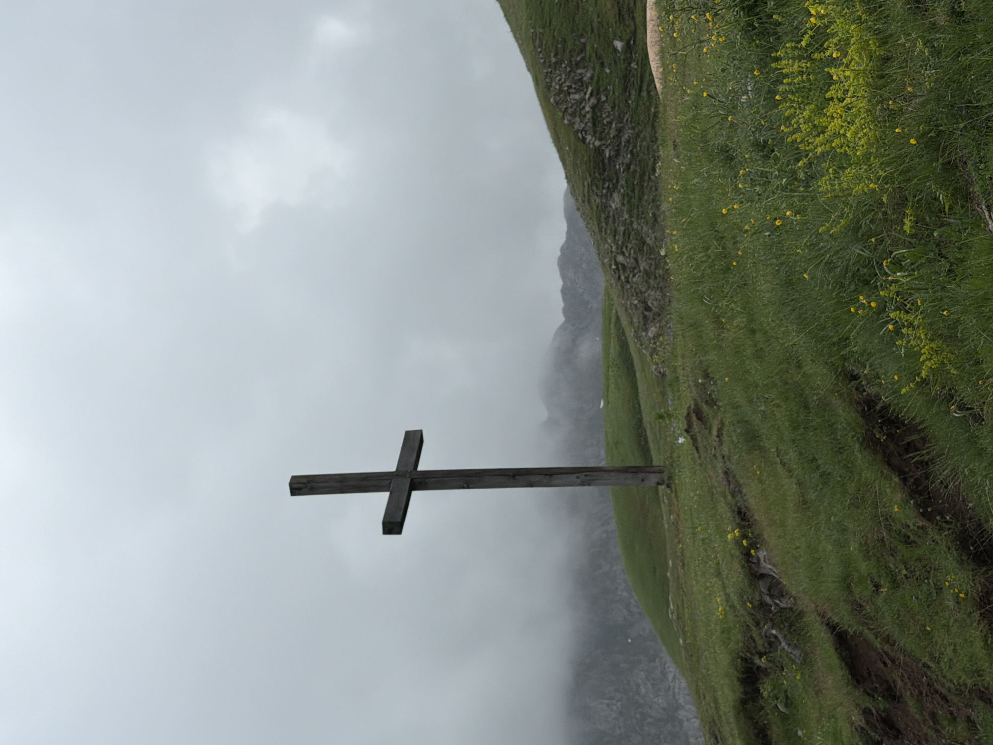 A solitary, weathered wooden cross stands upright on a verdant mountain slope, slightly to the left of the image's center when correctly oriented. The cross is made of simple, dark timber, with a thick vertical beam and a shorter horizontal crossbar.
The surrounding terrain is a lush green hillside, densely covered in grass and speckled with numerous small yellow wildflowers. Patches of rock outcrop are visible among the vegetation, hinting at the ruggedness of the mountainous environment. In the background, distant mountains are partially obscured by a thick, gray mist or low-hanging clouds, which blend seamlessly with the overcast sky. The sky itself is a uniform, light gray, indicating a cloudy day with diffuse light.
No people are visible, and there is no discernible activity taking place, conveying a sense of quietude and remoteness. The setting suggests a high-altitude location, fitting for Kapetanovo Jezero, Montenegro. There is no visible text on the cross or elsewhere in the image. The overall atmosphere is serene and somewhat solemn due to the misty conditions and the prominent cross.