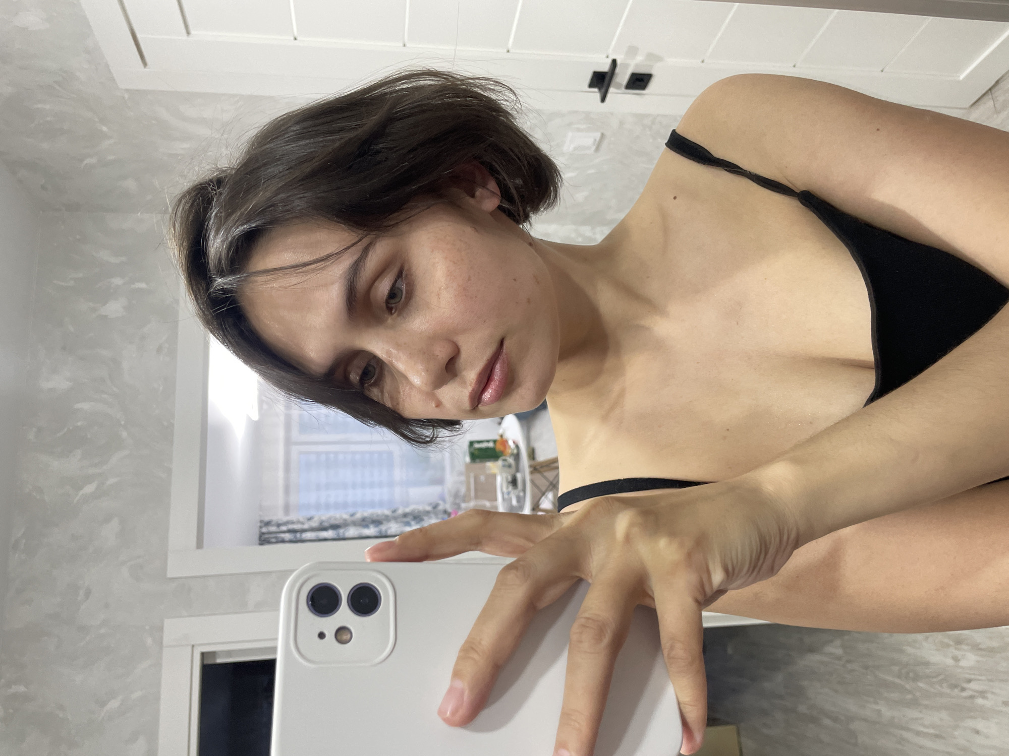 A woman with short brown hair and fair skin is taking a selfie in what appears to be a bathroom. She is wearing a black top. She is holding a white smartphone with two visible camera lenses. The background includes a marble-patterned wall, a white door with black hardware, and a window with blinds partially visible. The location is not apparent from the image. Therefore, there is no information available to confirm this photo was taken in Izhevsk, Russia.
