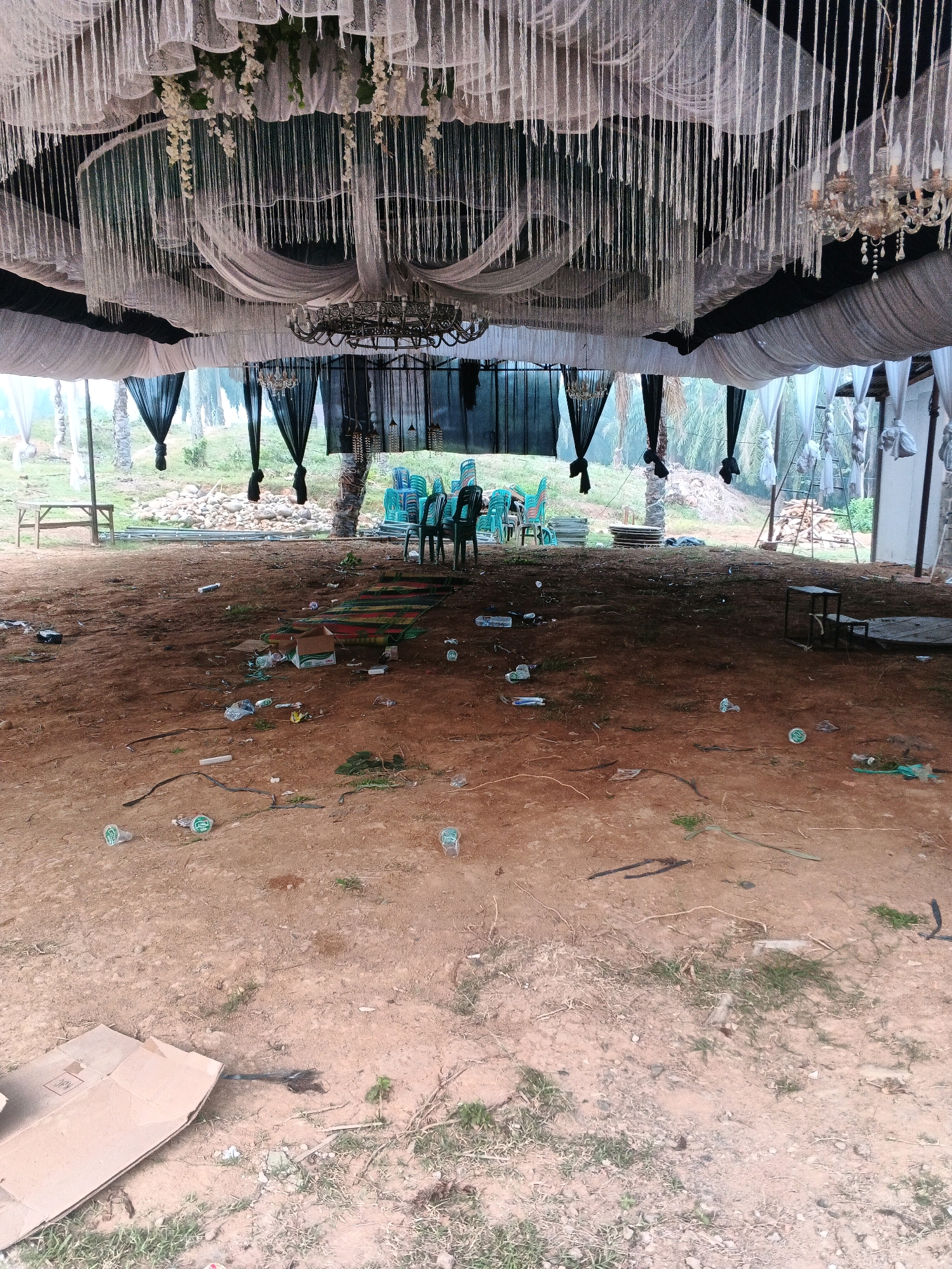 The image depicts an outdoor event venue in Brangan Mulya, Indonesia, that has been left in a state of disarray. The scene is under a tent or canopy decorated with draped white and black fabric, along with hanging strings of beads or similar material and chandeliers. The ground is dirt and appears to be littered with trash, including plastic bottles, wrappers, and a discarded cardboard box. In the background, stacks of green chairs are visible, suggesting they were used for seating. A mat or rug is also lying on the ground. The overall impression is of a recently concluded event that has not yet been cleaned up. The environment beyond the tent appears to be rural, with vegetation and trees visible.