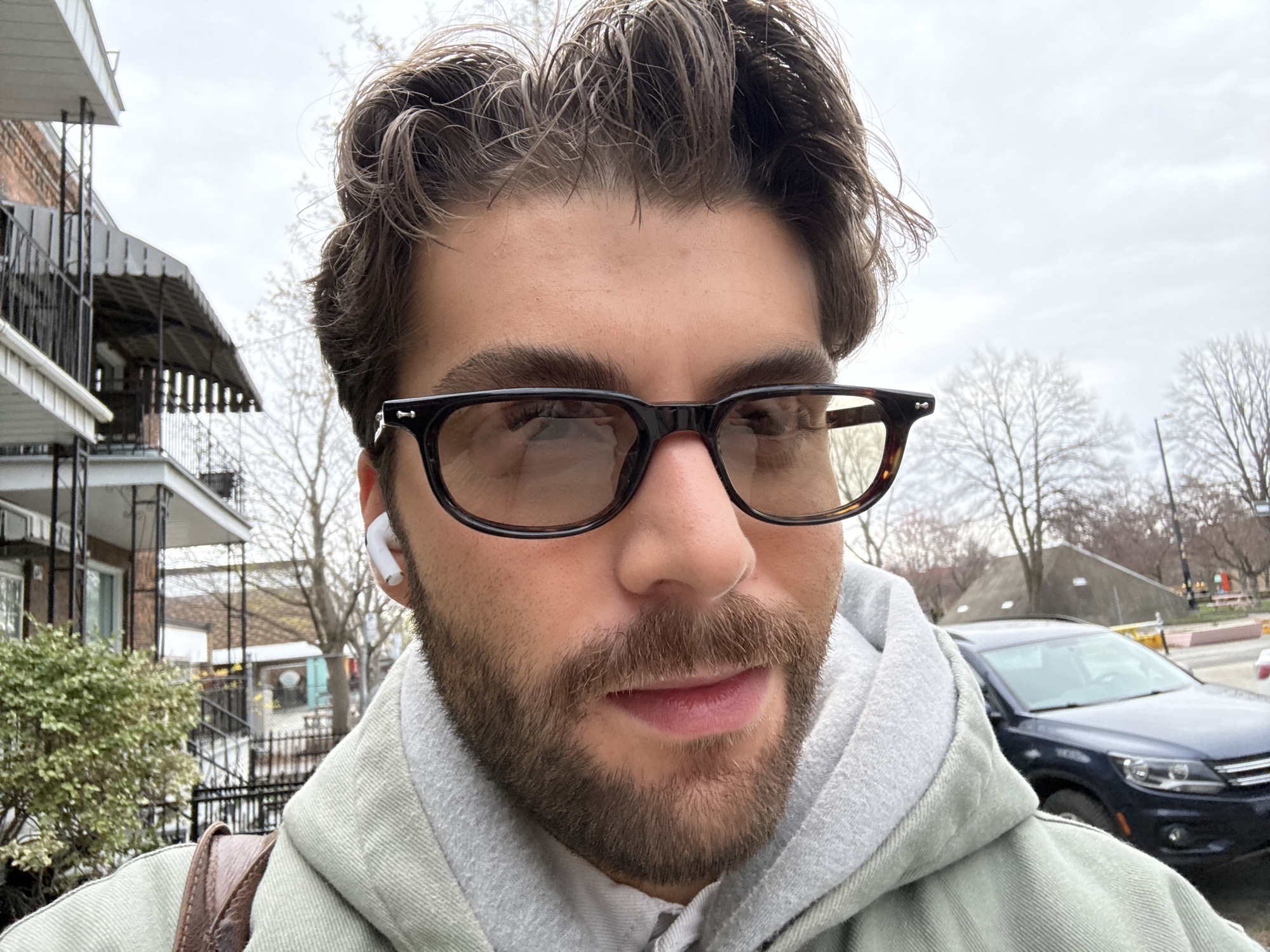 A close-up, selfie-style photograph captures a man outdoors on an overcast day, likely in Montreal. He looks directly at the camera with a neutral expression. The man has dark, medium-length wavy hair, prominent dark eyebrows, and a full dark brown beard and mustache. He wears dark-rimmed eyeglasses with subtly tinted lenses, and a white wireless earbud is visible in his right ear. His attire consists of a light grey hoodie under a light greenish-grey jacket, with a brown leather strap (likely from a bag) visible on his left shoulder.
The background on the left side shows residential buildings with balconies and dark awnings, alongside green bushes and bare trees. On the right, a dark blue SUV, appearing to be a Volkswagen Tiguan, is parked on the street, with more bare trees and a distant light post visible. The sky is uniformly grey and overcast, indicating a cloudy daytime setting. The bare trees suggest the photograph was taken during late winter or early spring in a temperate climate like Montreal. There is no visible text in the image.