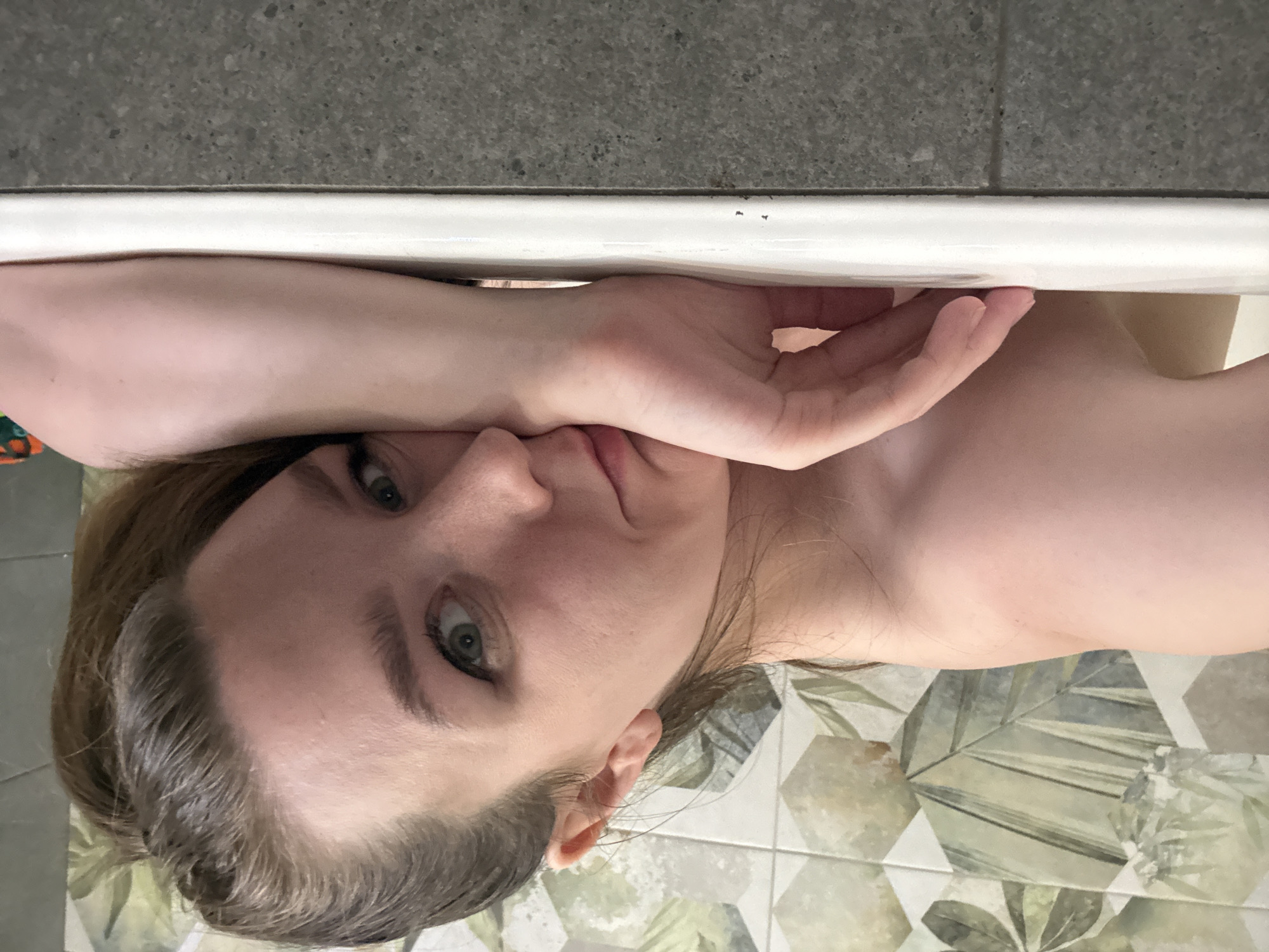A woman is lying in a bathtub. She is light-skinned with light brown hair. Her left arm is draped over the edge of the tub, her hand reaching over her face. The background consists of a gray wall, the white edge of the bathtub, and patterned floor tiles in shades of gray, green, and beige. The location is unknown, and there is no evidence indicating it is Izhevsk, Russia.