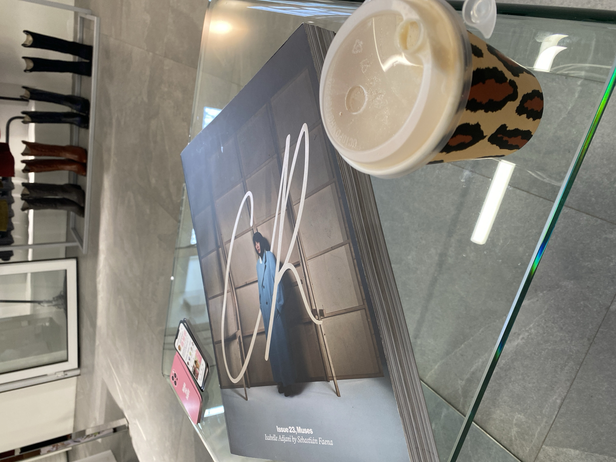 The image shows a glass table with a magazine, a coffee cup, and a pink smartphone. The magazine is titled "Issue 23, Muses Isabelle Adjani by Sebastián Faena." The coffee cup has a leopard print design. A pink smartphone is on the left side of the table, next to the magazine. The table appears to be in a retail environment, possibly a fashion boutique, as evidenced by the reflection of clothing racks and boots in the mirror in the background. The city is not discernible from the image.