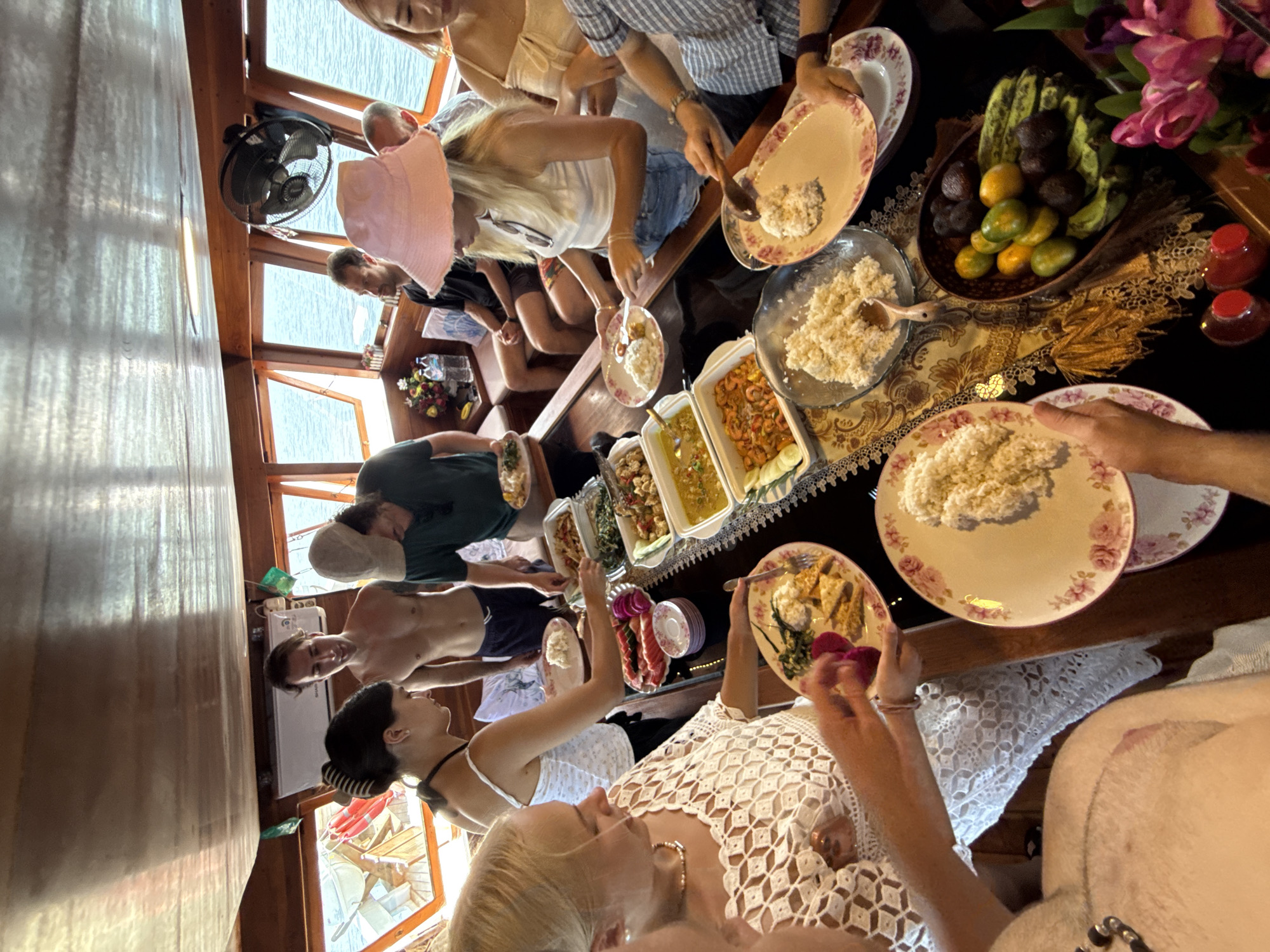 A group of people are gathered around a table laden with various dishes and bowls of food. The table is covered with a patterned tablecloth, and there are plates of rice, fruits, and other delicacies. The scene appears to be indoors, potentially on a boat, given the presence of a window with a view of the sea and the wooden paneling on the walls. The atmosphere suggests a gathering or meal shared among friends or family. The location is in the city of Unknown, Indonesia.