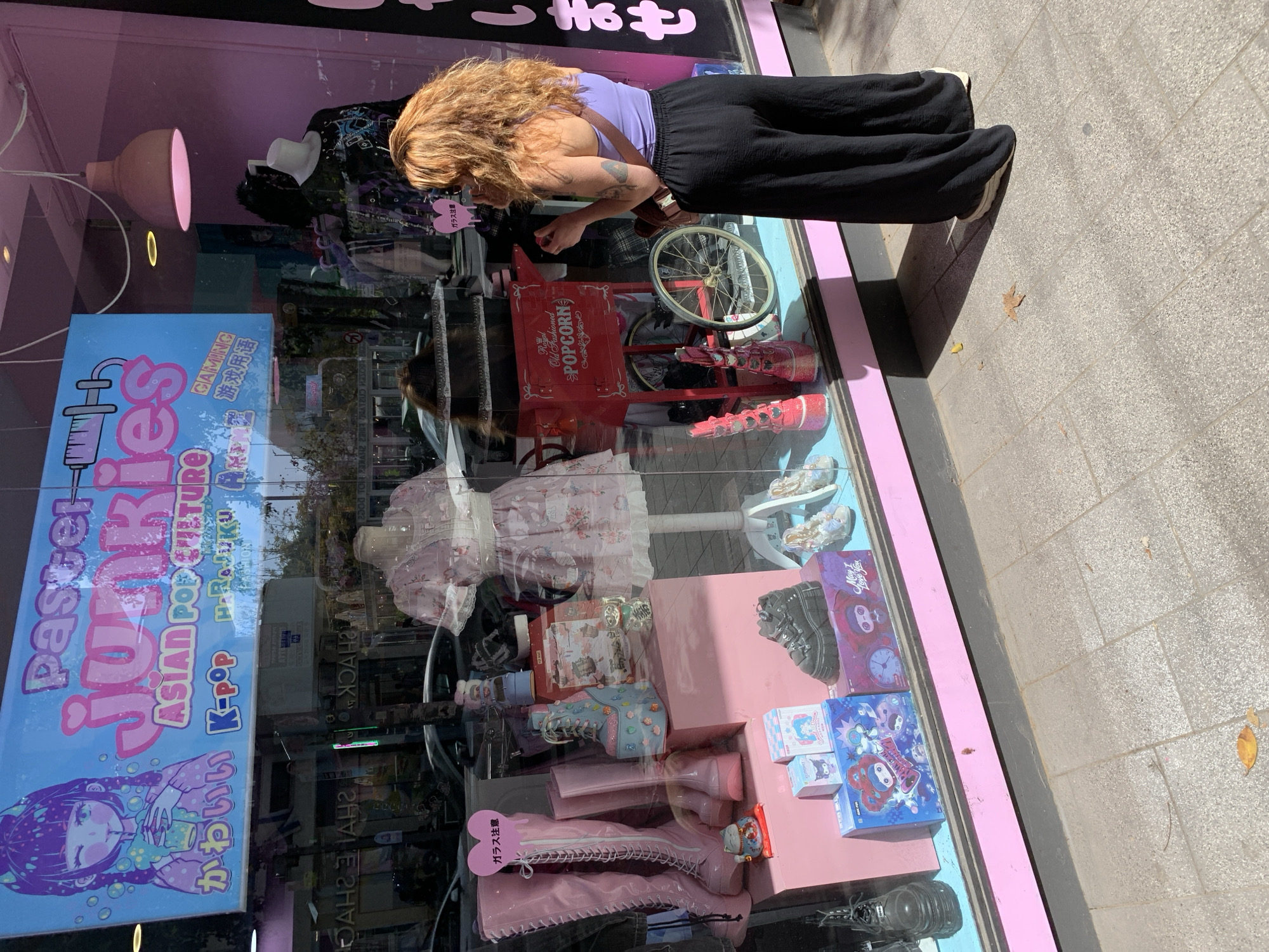 The image shows the storefront of "Pastel Junkies," an Asian pop culture store, presumably located in Tel Aviv, Israel, based on the Hebrew writing on the storefront. A woman with curly blonde hair, wearing a purple top and black pants, is looking into the store window. The store's display includes pink-themed items, such as boots, clothing, and figurines, along with a popcorn machine. The storefront also has signs in Japanese and English. The sidewalk is visible outside the store, with leaves scattered on the ground.