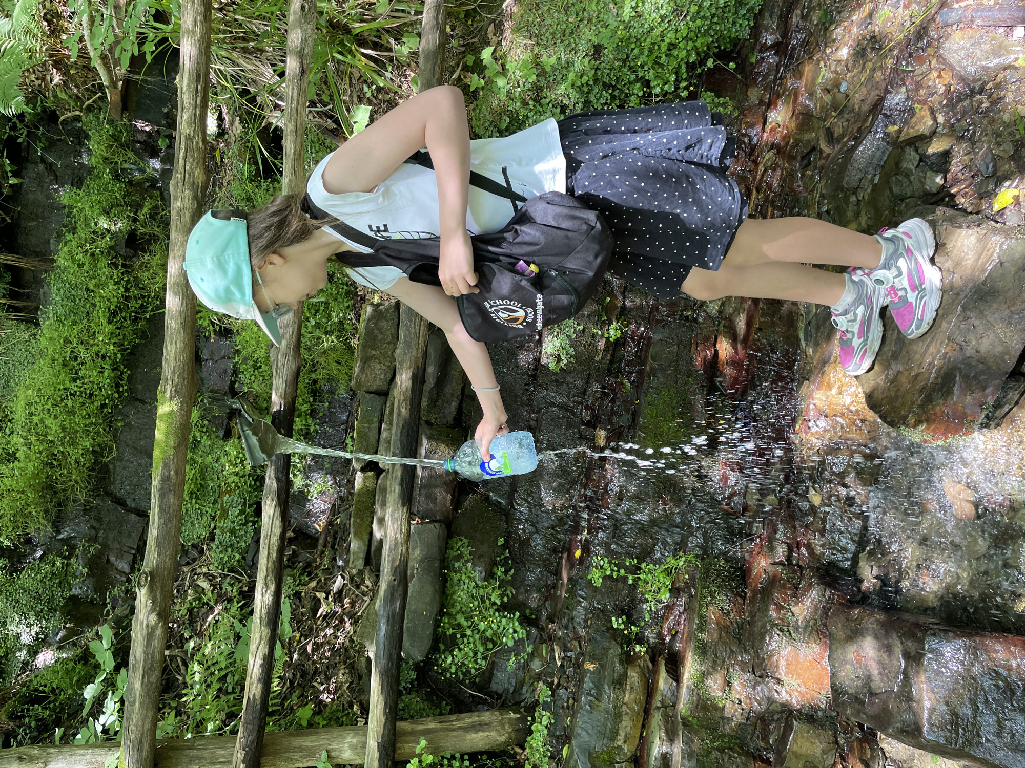 A person is pouring water from a plastic bottle into a stream in a wooded area. The person is wearing a white t-shirt, a black skirt with white polka dots, a light blue cap, and athletic shoes. They are also carrying a black backpack. The stream flows over rocks and is surrounded by lush green vegetation. The sunlight suggests it is daytime, and the weather appears to be pleasant. The scene has a natural and serene feel.
There is visible text on the backpack. One part of the text reads "SCHOOL" and another part appears to be "3000". The label on the water bottle also has some text but it is not clearly legible. The location is contextualized as Izmaylovka, Russia.