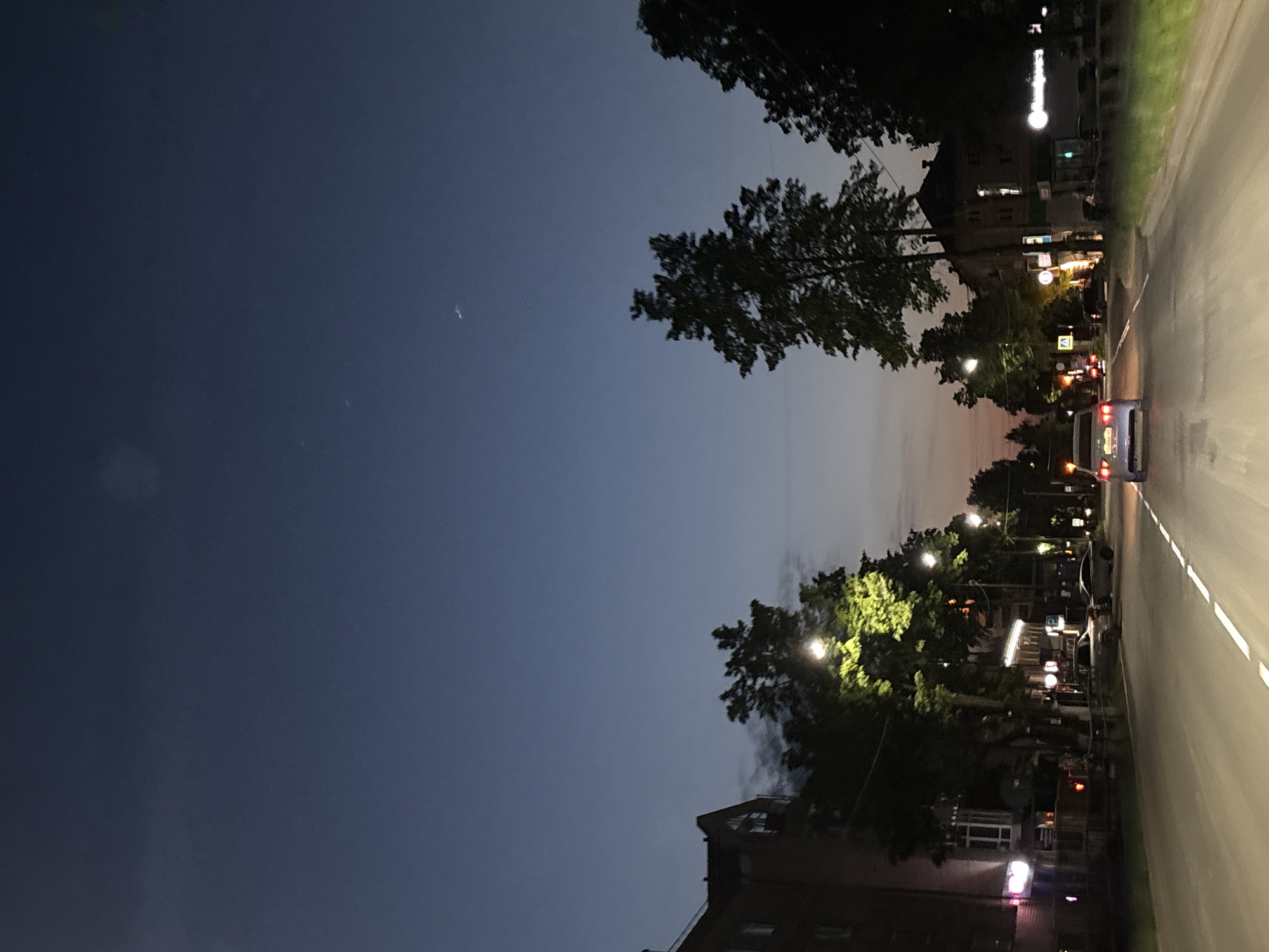 A nighttime street scene in Chaikovsky, Russia. A road stretches into the distance with a solid white line marking the edge. Cars are visible on the road, with one close enough to show taillights. Buildings line the road, interspersed with trees. Streetlights illuminate the trees, buildings, and roadway. The sky transitions from dark blue to a lighter hue near the horizon. A crescent moon is visible in the dark blue portion of the sky.