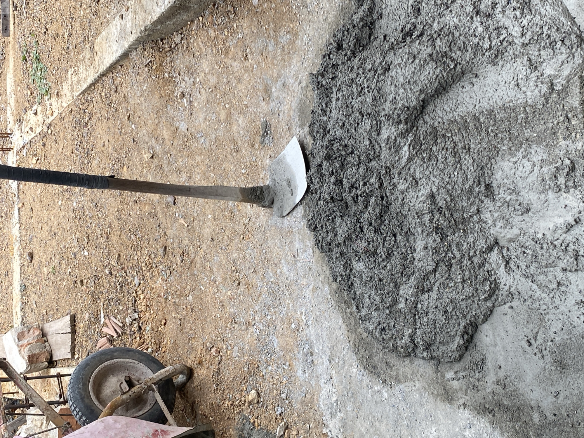 A shovel with a black taped handle rests in a pile of wet cement on a dirt and gravel ground. A wheelbarrow with a grey tire and a pink sheet is visible in the upper left corner. In the background, there are scattered rocks and a concrete curb. The scene appears to be a construction site.