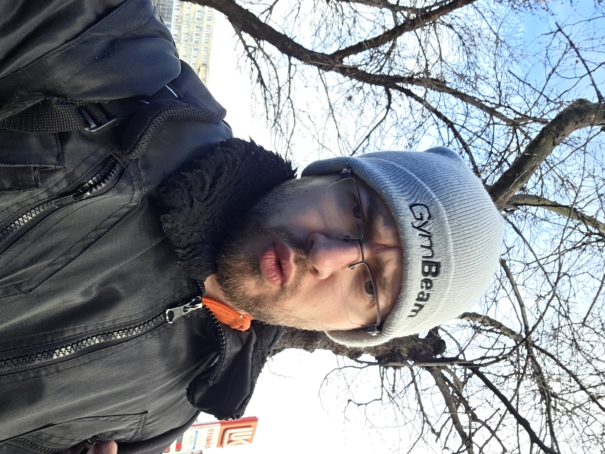 A man in a grey beanie labeled "GymBeam" and glasses is wearing a dark winter jacket with a fur collar and an orange accent. He appears to be standing outdoors on a partly sunny day with bare tree branches visible in the background. A building is visible in the far background. A sign with Cyrillic text is partially visible in the lower-left corner of the image. The image appears to have been taken at an upward angle. Based on the sign in the lower-left, the image was taken in the city of Plovdiv, Bulgaria.