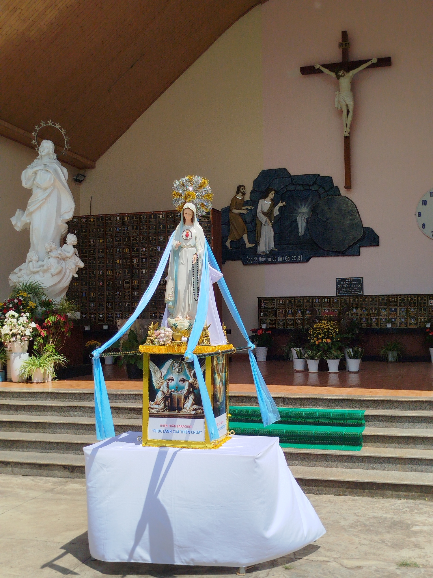 The image shows a Catholic church exterior in Pleiku Ward, Vietnam. In the background, to the left, stands a white statue of the Virgin Mary with a halo. Adjacent to the statue is a flower arrangement on the same level. To the right, a crucifix is mounted on the wall. Below the crucifix, a depiction of the empty tomb of Jesus is sculpted onto the wall. In front of the wall are several rows of memorial plaques and plants in white pots. In the foreground, a table draped with a white cloth holds a statue of the Virgin Mary adorned with a golden crown and blue fabric. The base of the statue features a golden frame displaying an image of angels and text in Vietnamese. The scene suggests a religious celebration or dedication. The location is recognizable from the Vietnamese text and the ward.