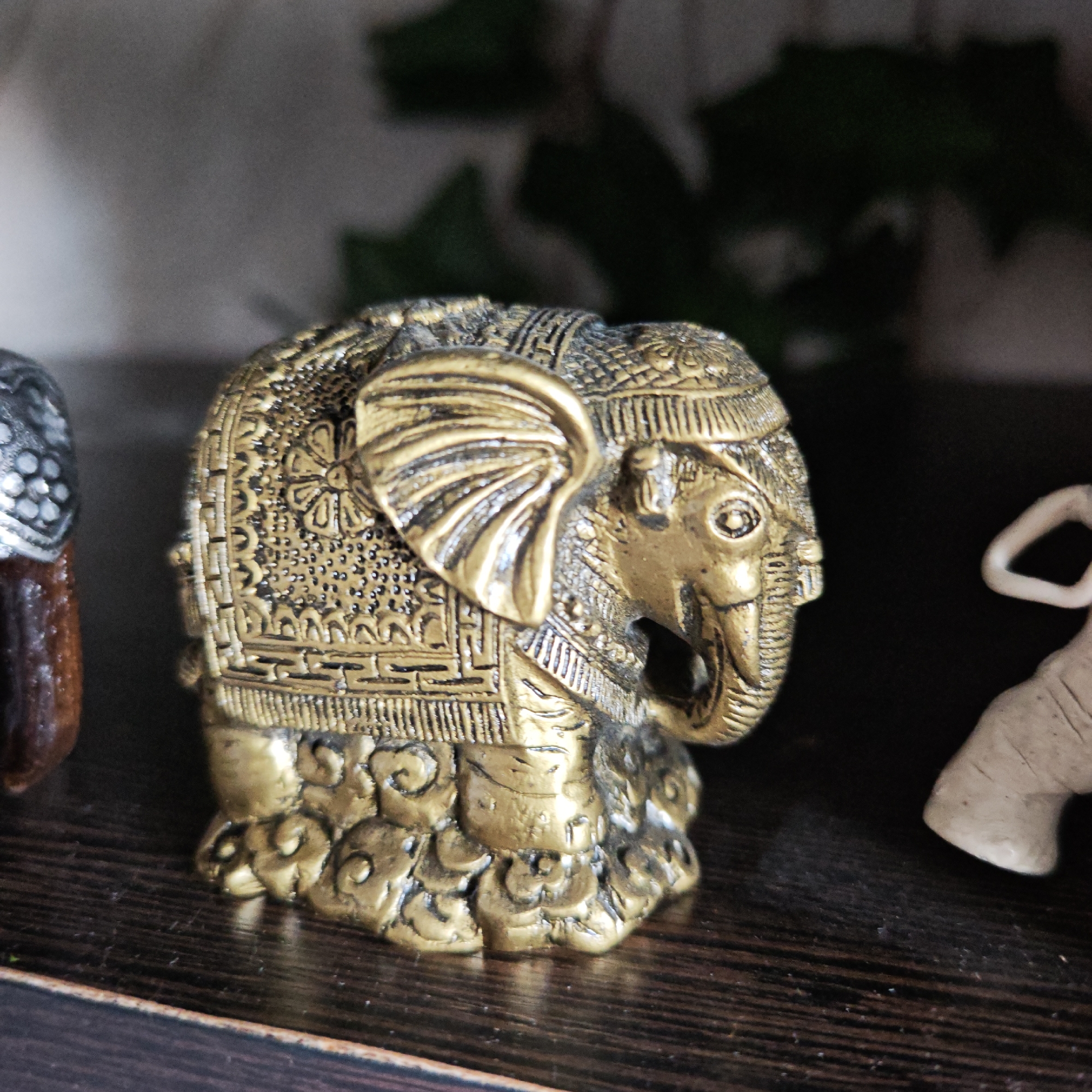 The image shows a golden elephant figurine with ornate detailing. The figurine is positioned on a dark wood shelf, with part of another figurine visible to the left. There is a vague shape of what may be foliage in the background. There are no people visible. Location information is not discernible from the image.