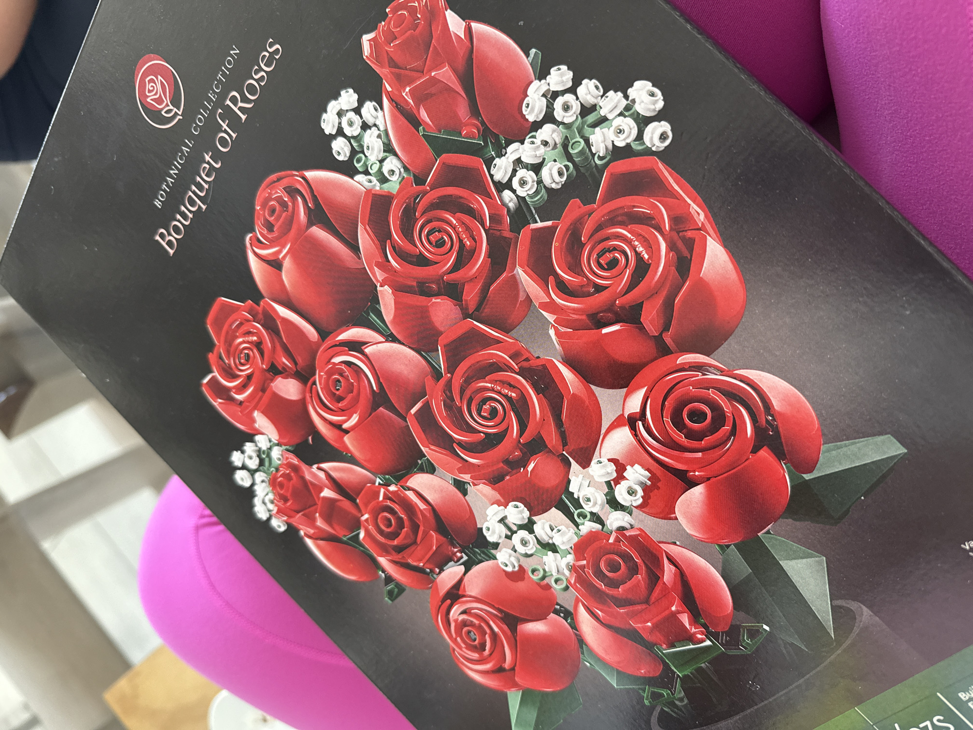 The image shows a close-up of the packaging for a Lego Botanical Collection Bouquet of Roses. The box is black with a picture of a bouquet of red roses with white accents. The words "BOTANICAL COLLECTION" and "Bouquet of Roses" are printed near the top of the box. Part of a person wearing bright pink leggings is visible in the background. The location is not discernible from the image.