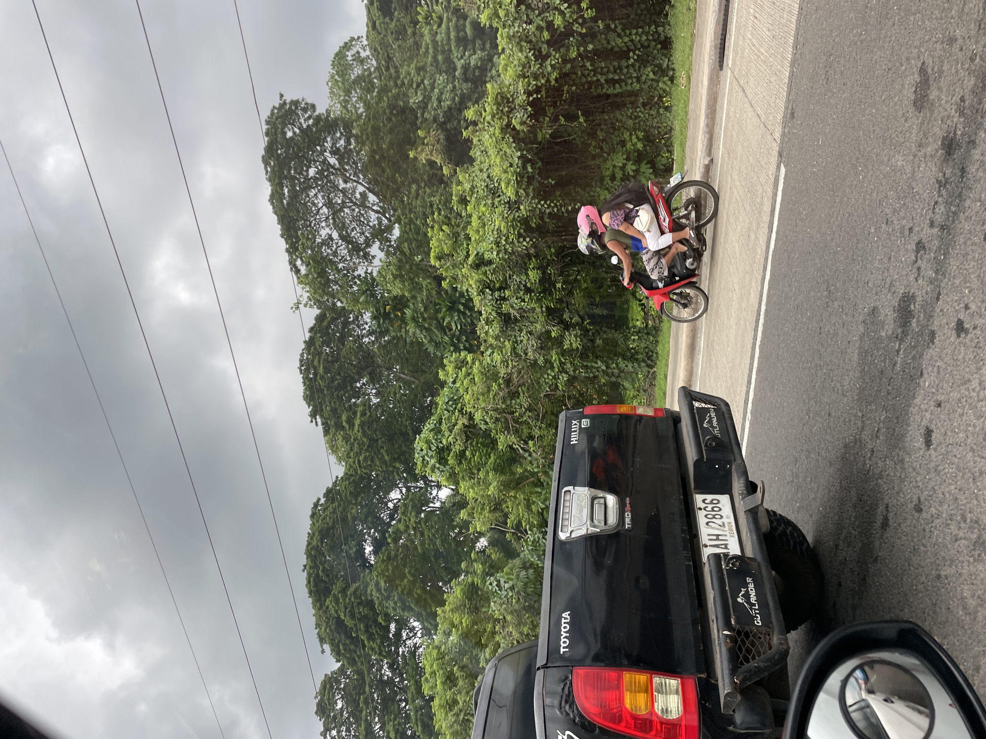 The image depicts a scene on a road in Davao City, Philippines. A black Toyota Hilux pickup truck is in the foreground, with a motorcycle carrying two people on the shoulder of the road. Both riders are wearing helmets, with the passenger's helmet being pink. Lush green trees line the roadside, and power lines are visible against a cloudy sky. The license plate on the Hilux reads "AH 2866."