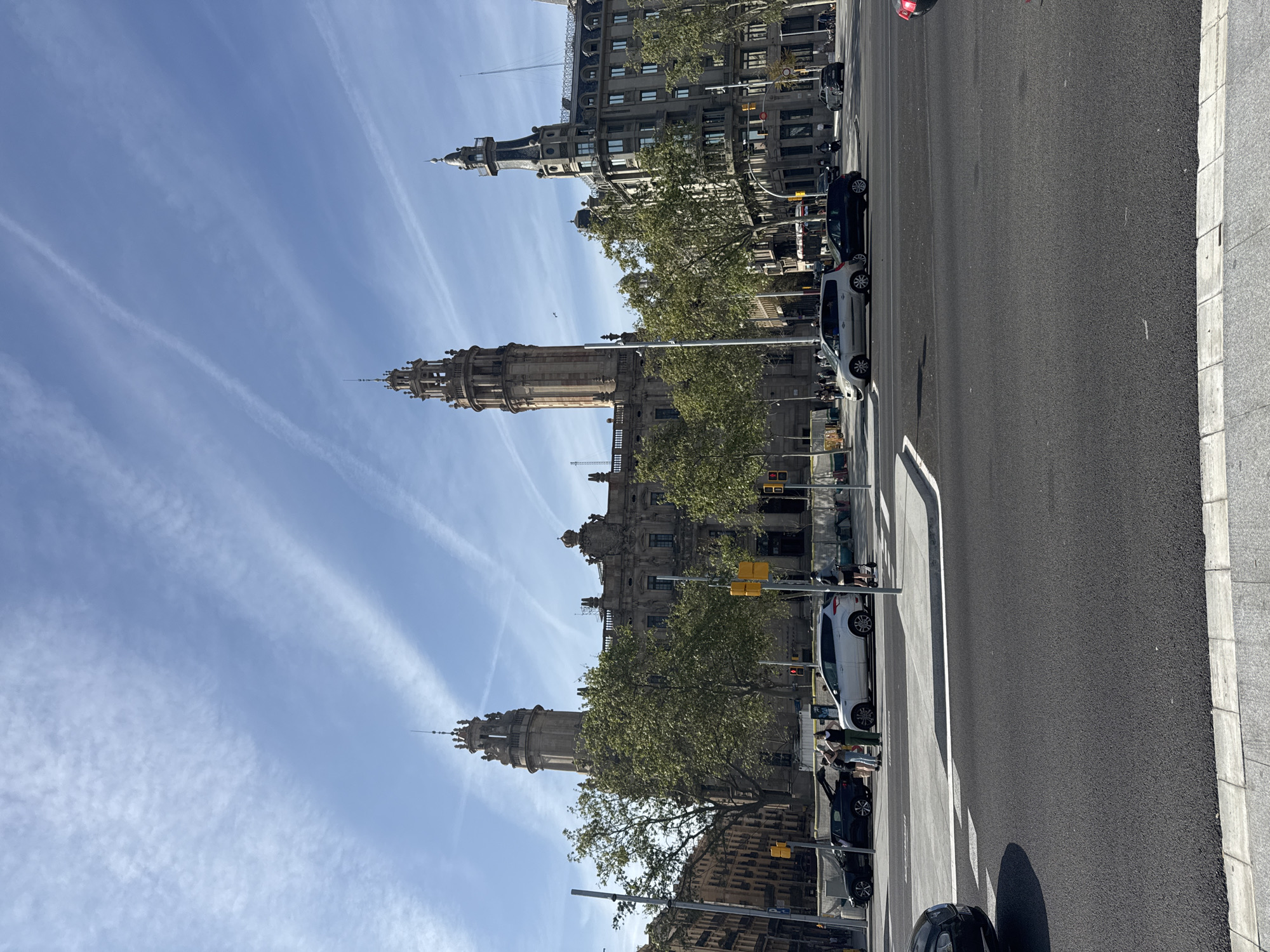 The image captures a street scene in Barcelona, Spain. The Passeig de Colom, a major thoroughfare, is depicted, lined with buildings of varying architectural styles and heights. The prominent landmark in the background is the Basilica de la Mercè, distinguished by its ornate facade and steeple.
Several cars are parked along the right side of the street. A few pedestrians stand near the parked vehicles. The sky is bright blue with wispy clouds. The street itself is paved with asphalt.