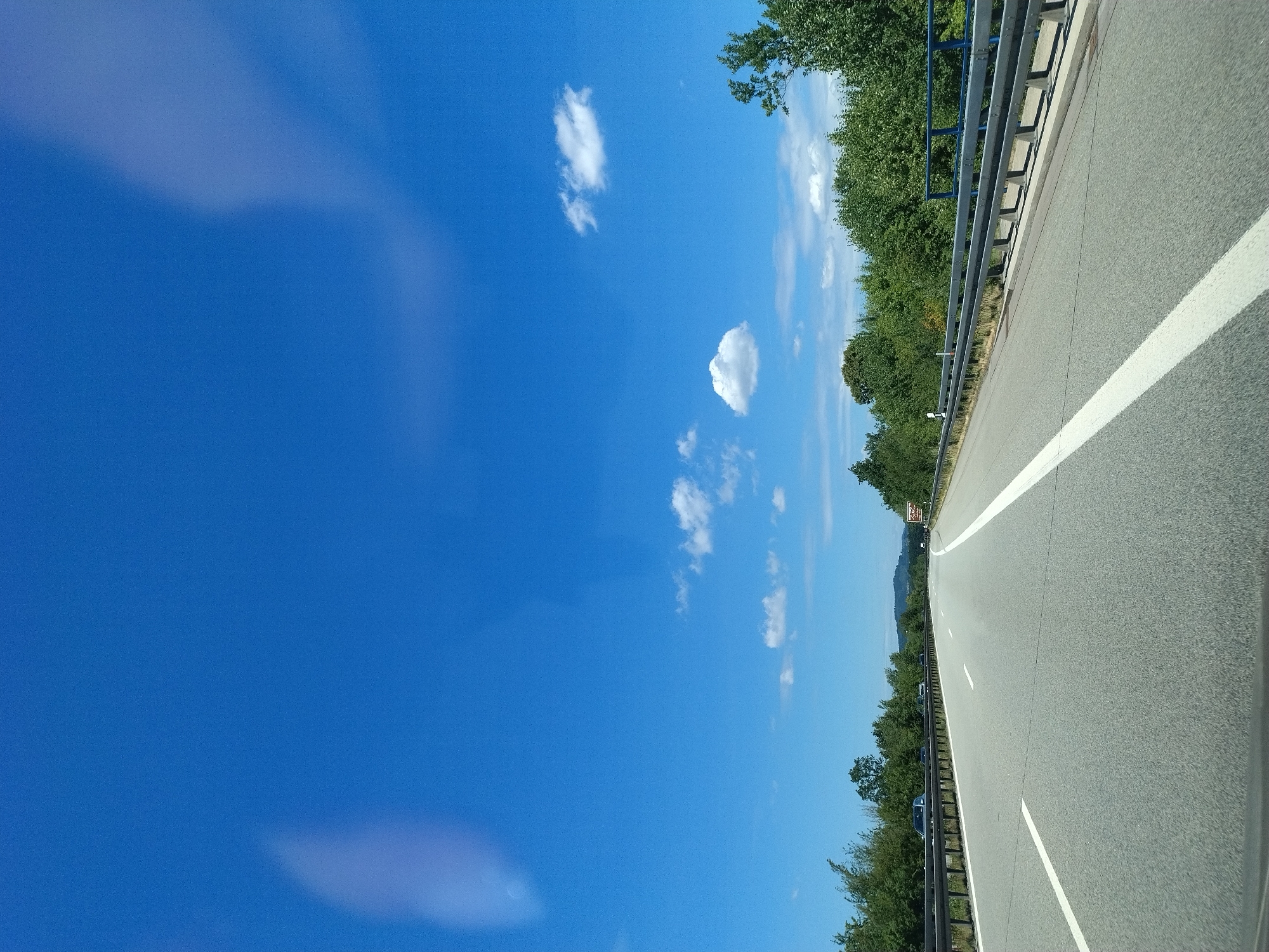 The image is a highway scene captured from the perspective of a vehicle in motion on a clear, sunny day. The sky is a vibrant blue with scattered white clouds. The highway is paved and marked with white lane lines. A metal guardrail runs along the right side of the road. Dense green trees and foliage line the sides of the highway. In the distance, a gentle hill with trees is visible. There are several cars on the highway, all appearing to be driving in the same direction as the viewpoint. The scene is devoid of people. The image is taken in Niederwinkling, Germany.