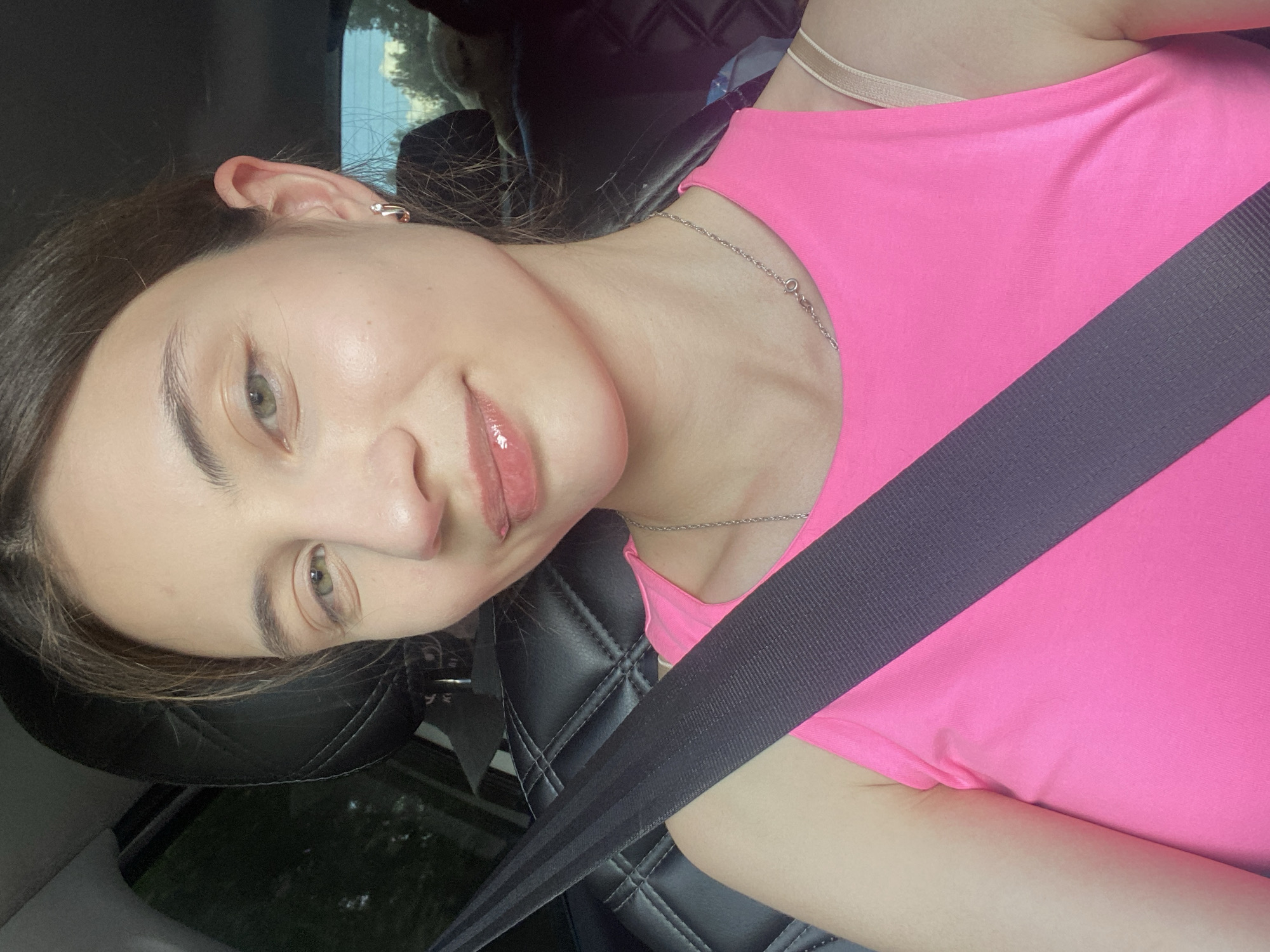 A young woman with light skin, brown hair, and green eyes is seen from the chest up, inside a car. She is wearing a bright pink tank top and a silver necklace. A black seatbelt is prominently displayed across her chest. Her lips are glossy and slightly parted, and she is looking directly at the camera with a subtle smile. The background shows the interior of a car, with a black leather seat and a glimpse of trees and sky through the window. No specific location within the city of Kamkoye, Russia, can be determined from the image.