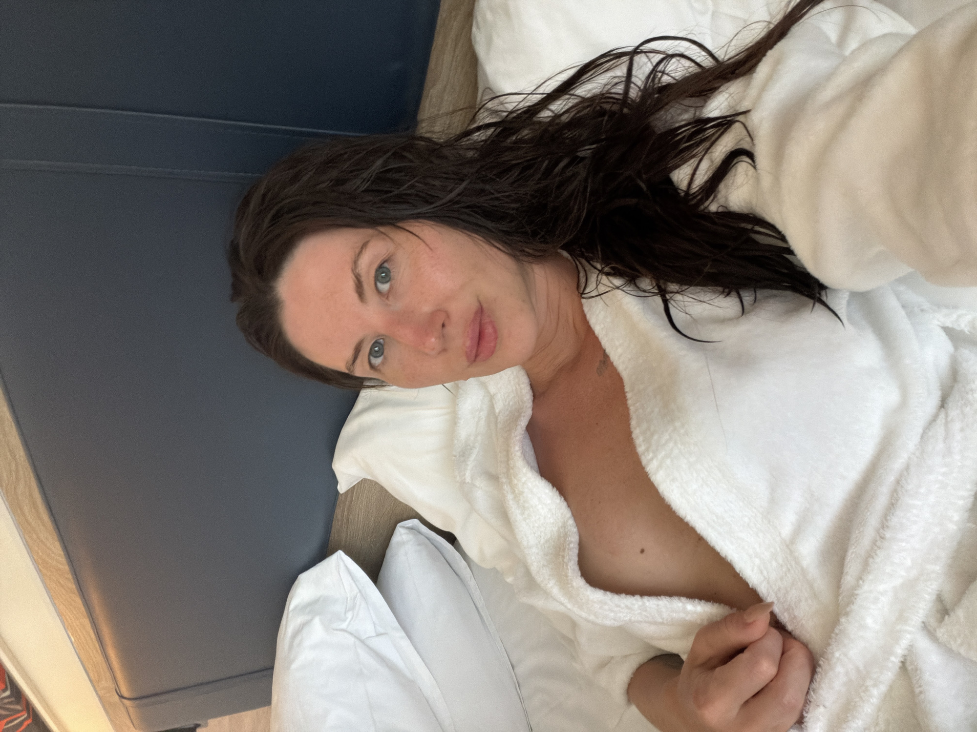 A woman with long, dark, wet hair is lying in a bed wearing a white bathrobe, which is open, revealing part of her chest. She is looking at the camera with a neutral expression. The bed has white pillows and a dark blue headboard. The background is blurred, but appears to be a room. The scene is indoors, and the lighting suggests it is daytime. The woman is positioned in the center of the frame. The image is a selfie, taken from a slightly high angle.
