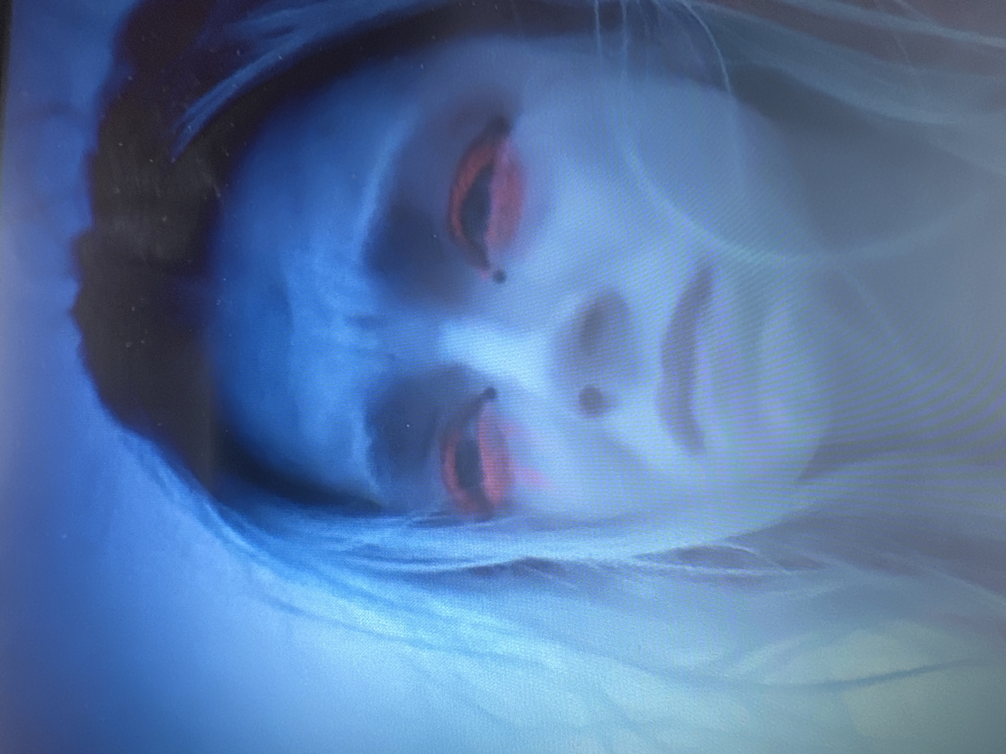 The image shows a close-up of a person's face, heavily stylized with bright blue skin and dramatic red eyeshadow. The person appears to be looking upwards with a distressed expression, their eyebrows furrowed. There are small dark dots on their cheeks, possibly beauty marks or part of the makeup design. The lighting is predominantly blue, creating a surreal and otherworldly atmosphere. The background is out of focus, appearing as streaks of blue and dark tones, suggesting it could be a screen display or a very dimly lit environment. There is no information available in the image to indicate a location in Plovdiv, Bulgaria.