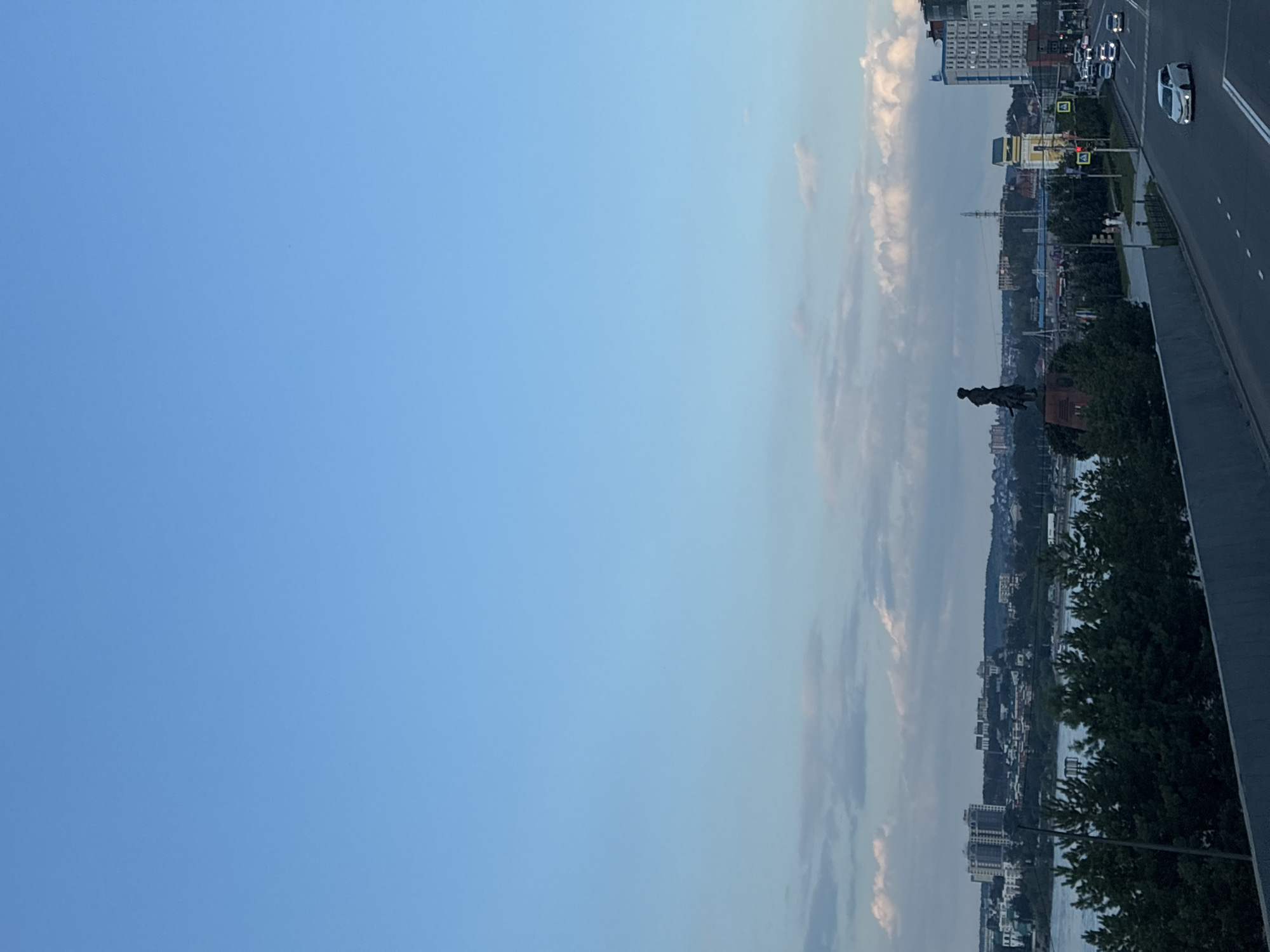 The image shows a cityscape in Irkutsk, Russia, during what appears to be late afternoon or early evening. A wide river, likely the Angara River, dominates the lower half of the scene, with buildings and trees lining its opposite bank. In the foreground, a highway runs diagonally from the bottom right towards the left, with several cars visible.
A prominent statue of a man on horseback, a historical monument, stands near the riverbank on the far side of the road. To the right of the statue, a distinctive yellow and white tower with a clock is visible, and further to the right, a tall, modern white building stands out. The sky is a clear blue with scattered white clouds. Green trees, likely pine trees, are densely planted along the riverbank in the mid-ground, creating a natural barrier between the water and the urban landscape. The overall impression is one of a dynamic urban environment situated alongside a natural river and green spaces.