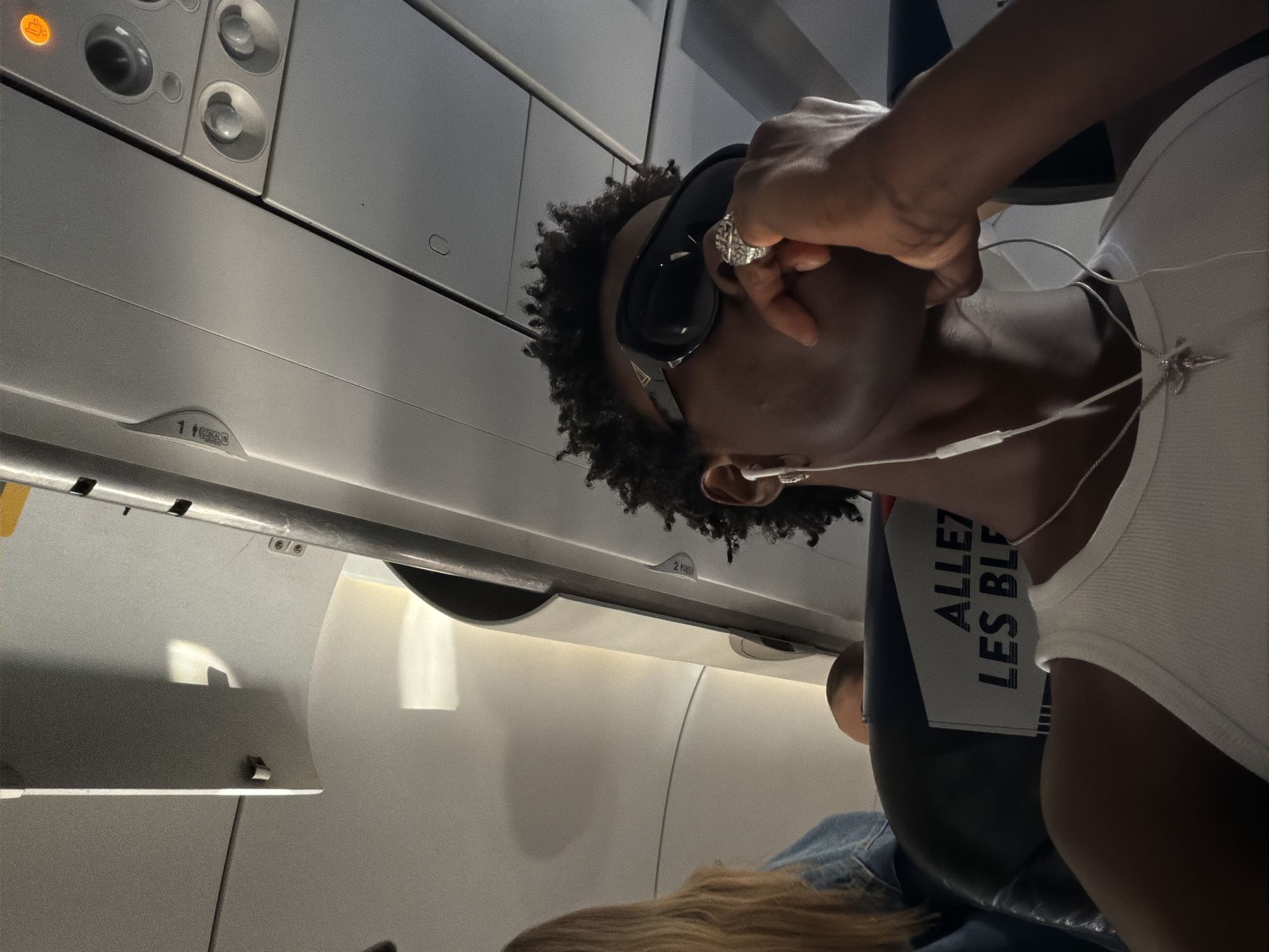 A person with dark skin, curly black hair, and wearing sunglasses is seated inside an airplane. The person is wearing a white tank top and a silver necklace. They have a ring on their finger and are holding the sunglasses. White earphones are visible. The interior of the airplane includes overhead compartments with safety labels and individual light and air controls. The view includes the heads of other passengers and part of their clothing. The person is sitting in an Allez Les Bleus seat. The image does not have anything to imply the location is Crawley, United Kingdom.