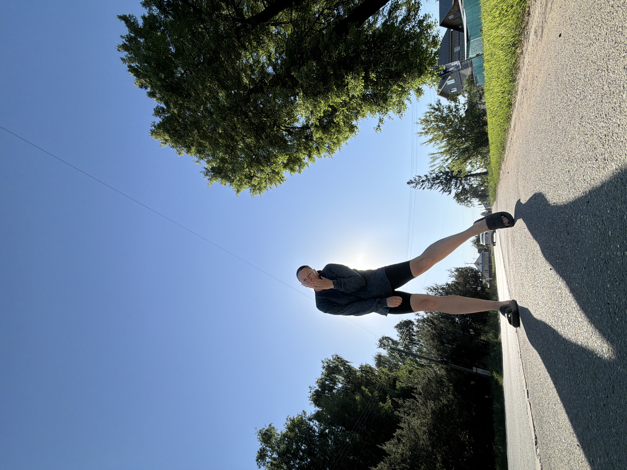 A person stands on an asphalt road under a blue sky in a location identified as Chaikovsky, Russia. They are wearing a long-sleeved gray shirt, black shorts, and dark sandals. Their hand is near their mouth. The sky is clear with the sun in the center of the image, and a tree is visible in the upper left corner. Other trees line the road, and buildings are seen in the background on the right side. An overhead wire is also visible in the sky. The image appears to be rotated counter-clockwise.