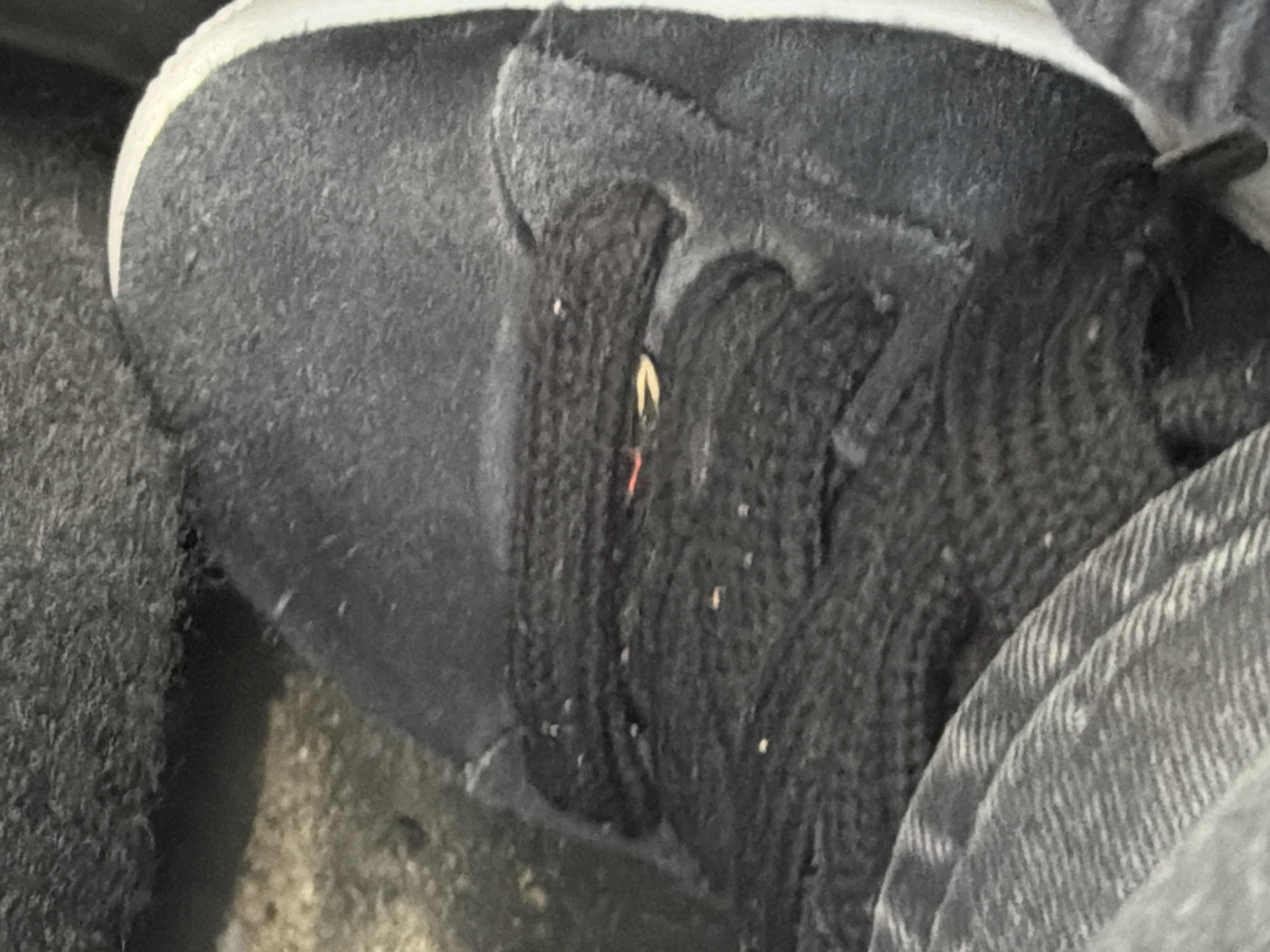 The image shows a close-up view of the top of a dark-colored shoe with black laces. The shoe appears to be made of suede and has a white accent line along the edge. The laces are thick and knotted. There are some small white specks on the laces and the shoe, possibly dust or debris. A small red and yellow object is visible between the laces, possibly a piece of string or a small foreign object. The background is dark and textured, suggesting an outdoor or industrial setting. The shoe is likely located on a street or pathway in Vallensbæk Landsby, Denmark.