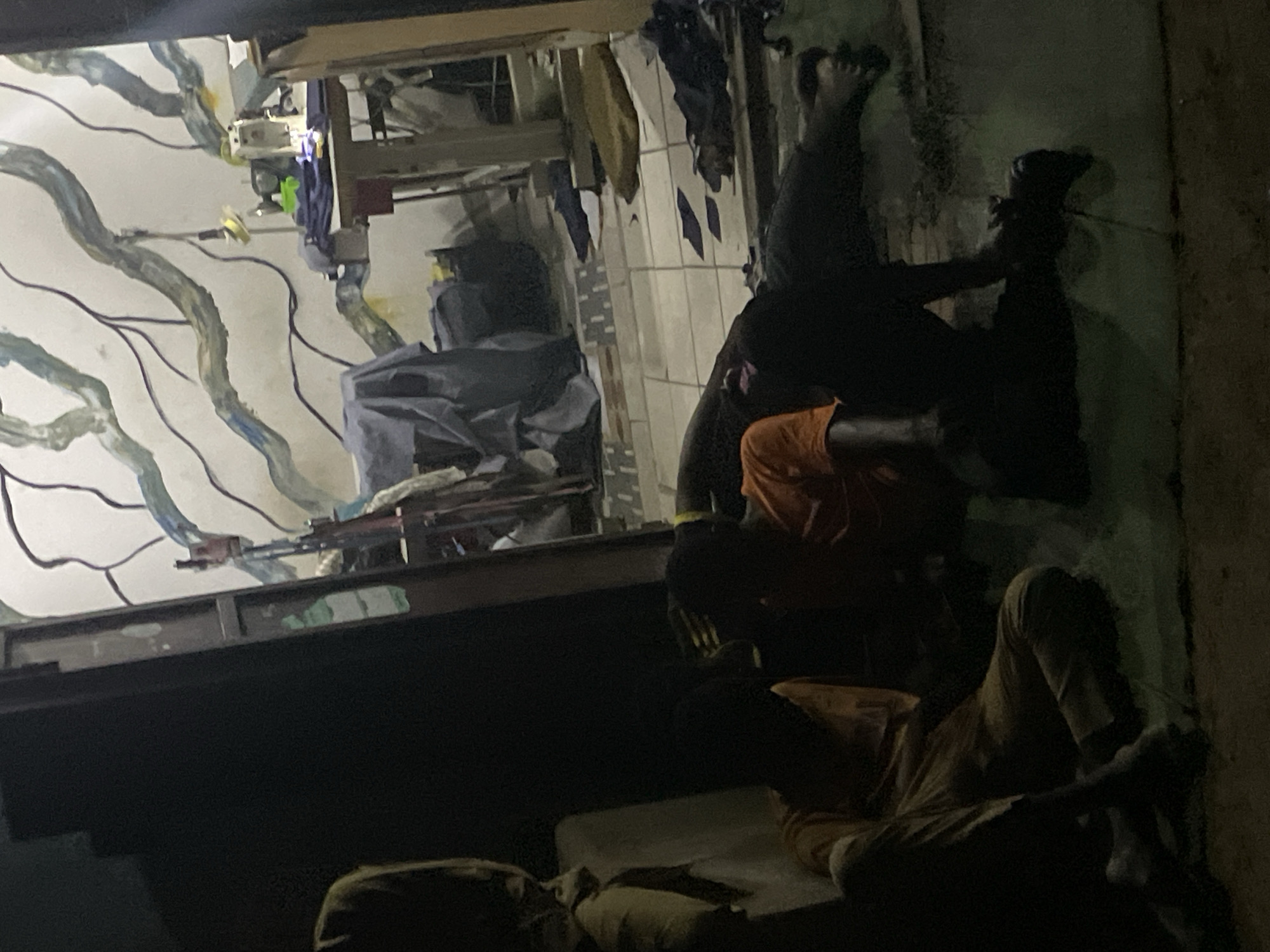 The photograph appears to show a dimly lit interior scene with a complex arrangement of objects and individuals. The setting seems to be a room or space with a distinctive wall pattern featuring light-colored lines or patterns against a darker background. The interior contains various items, including what appears to be clothing, bedding, and other miscellaneous objects.
Several individuals are visible within the scene. At least three people are distinguishable, each in varying poses. One figure is positioned near the wall with their feet raised, suggesting a leaning or resting posture. Another individual is seen in an orange shirt, while a third figure is lying down on what appears to be a mattress or bedding.
The environment is dark, and the image quality is somewhat grainy, which makes it difficult to discern finer details. The location is listed as Unknown, Unknown, so no specific details about the city or region can be determined. The overall impression is one of an informal, cluttered, and dimly lit living space.
