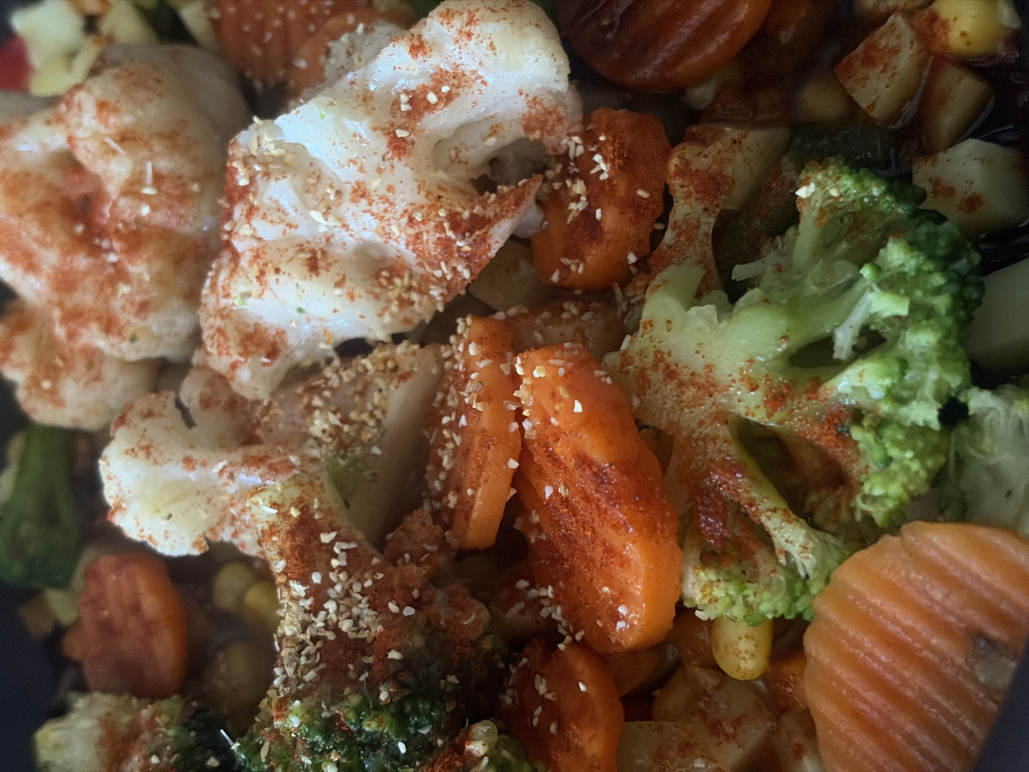 The image shows a close-up view of cooked vegetables, including cauliflower, broccoli, and sliced carrots. The vegetables are sprinkled with a red spice, possibly paprika, and small white seeds. The vegetables appear to be in a bowl or container. There are no people visible in the image. The location is not possible to determine from the image.