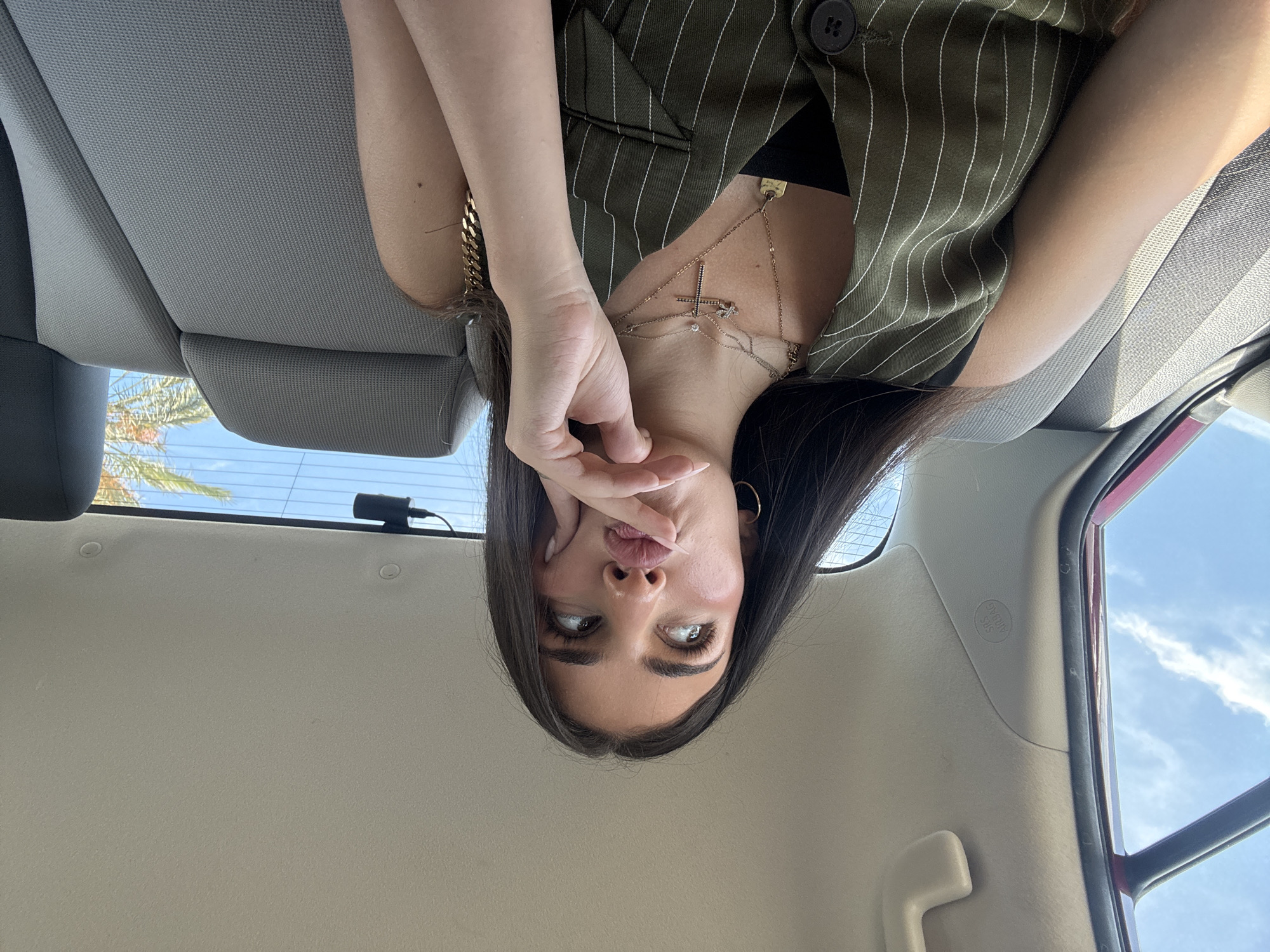 This is a slightly rotated, head-on selfie taken inside a car. The subject is a young woman with long, dark hair. She is wearing a green pinstripe vest over a black top and several necklaces, including a cross pendant. Her right hand is raised, with her fingers touching her mouth and chin, and she is making a "kissing" face. Her eyes are wide open and looking to her right.
The background shows the interior of the car, with gray fabric seats and headliner. Through the rear window, a bright blue sky with some white clouds and palm trees can be seen, suggesting it is daytime and likely warm weather. A small black device, possibly a dashcam, is visible on the top edge of the rear window. The overall impression is a candid, playful moment captured while traveling. The visible text "SRS AIRBAG" is present on a label inside the car.