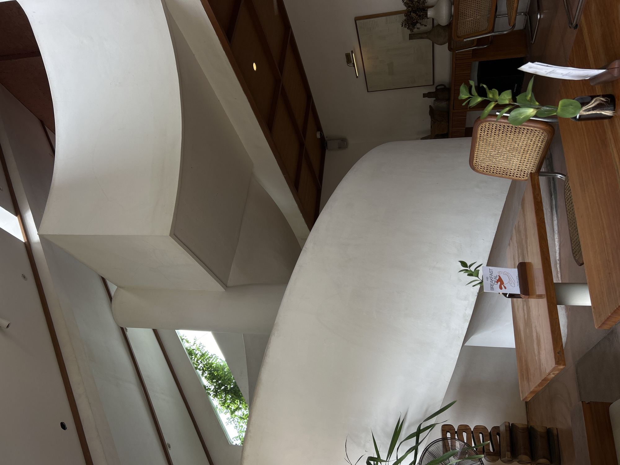 The image depicts an interior in Ubud District, Indonesia, featuring a distinctive architectural design with a curving white structure that appears to be a wall or staircase. Natural light floods in through a window showing greenery outside. A dining area is furnished with wooden tables and chairs with woven seats. There is a small plant in a vase on the table, and a menu card is placed on the edge of the table. The ceiling has wooden panels.