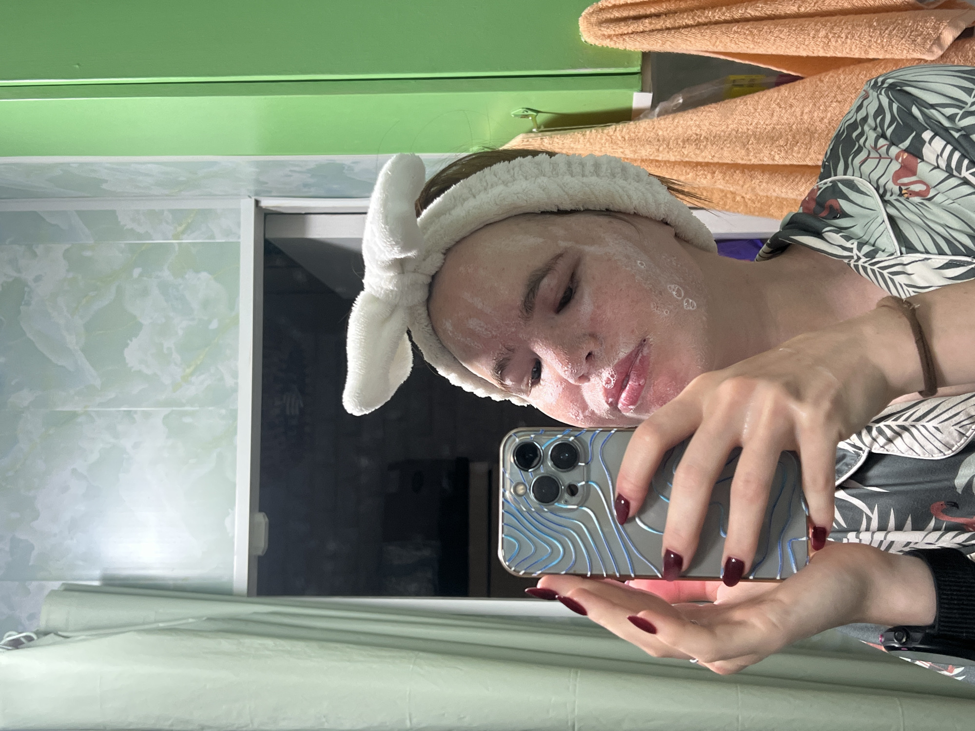 A young person with a white bunny-ear headband and a face mask is taking a selfie in a bathroom. The person is wearing a dark grey pajama top with a tropical print featuring flamingos and palm leaves. Their nails are painted a dark red color. The bathroom features light green, marbled wallpaper and a bright green cabinet. Two orange towels hang on a rack to the right of the cabinet. A light green shower curtain is visible on the left side of the image. The scene appears to be indoors, likely in a residential bathroom. The location of Chaikovsky, Russia is not discernible from the visual information provided in the image.