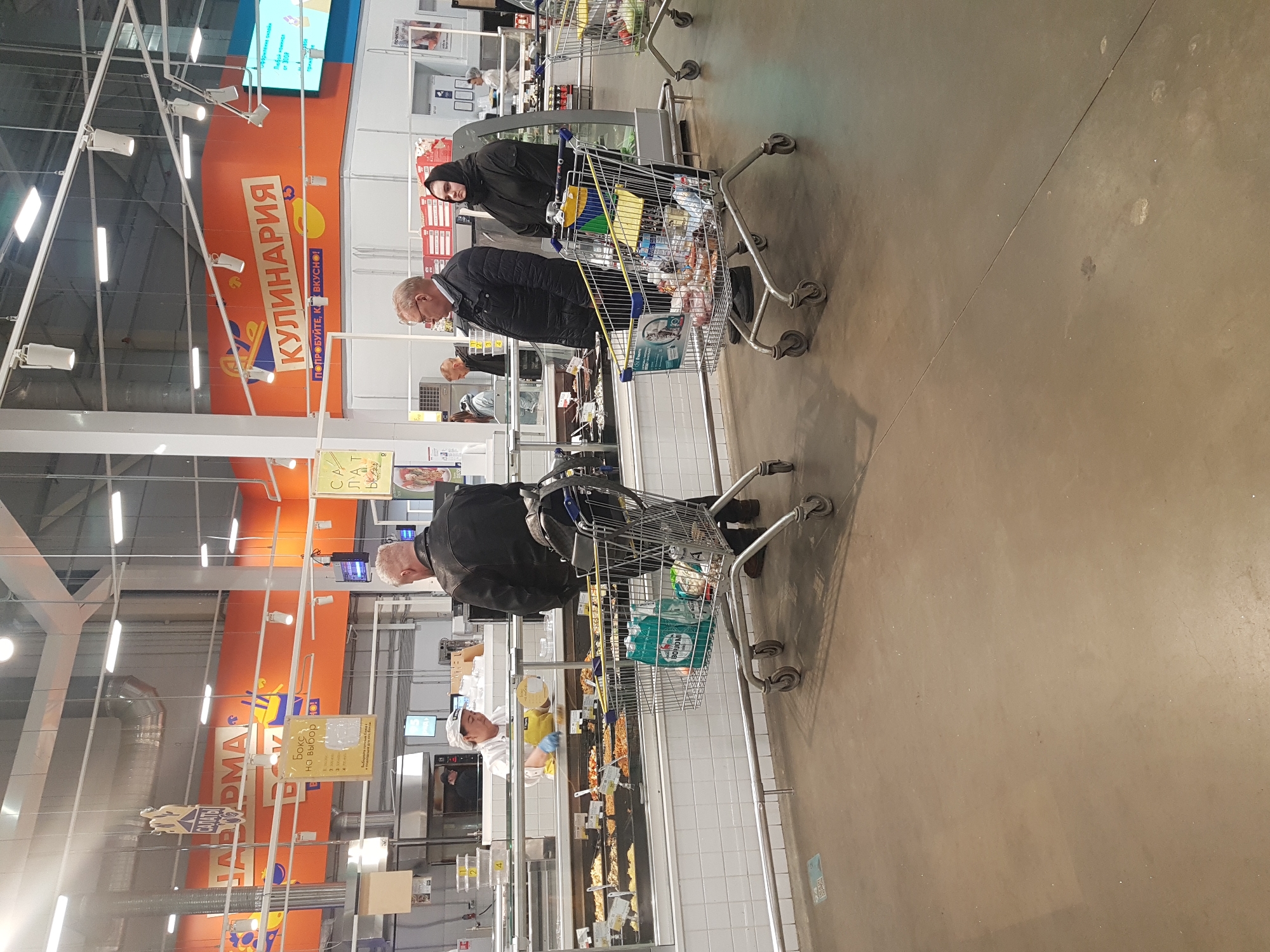 Here is a detailed description of the image:
**Scene:** The image depicts the interior of a supermarket or large grocery store. There are at least four customers visible with shopping carts in the foreground and middle ground.
**People:** The individuals are dressed in casual outerwear, including jackets and sweaters. One person is wearing a head covering. The people are likely customers selecting food items. One person in the middle ground appears to be a store worker, wearing a white hat and working behind a food counter.
**Objects:** There are several shopping carts containing a variety of food items. The store is well-stocked with various goods on display. There are overhead signs written in Russian, including the words "КУЛИНАРИЯ" (culinary), "CAЛАТЫ" (salads), and "Бокс на выбор" (box to choose from). Product packaging in the shopping carts indicates that some items are from recognizable brands.
**Actions/Interactions:** The customers are engaged in shopping activities, browsing and selecting food items from a refrigerated display counter. There are no overt interactions among the shoppers, as each seems focused on their own selection process.
**Environment:** The lighting is bright and seems to be typical of a retail environment. The overall setting is that of a bustling marketplace.
**Possible Location:** Given the Cyrillic text on the signage, the image is almost certainly from a store in Russia. It may be located in Saint Petersburg, Russia, if the photographer was in that location.