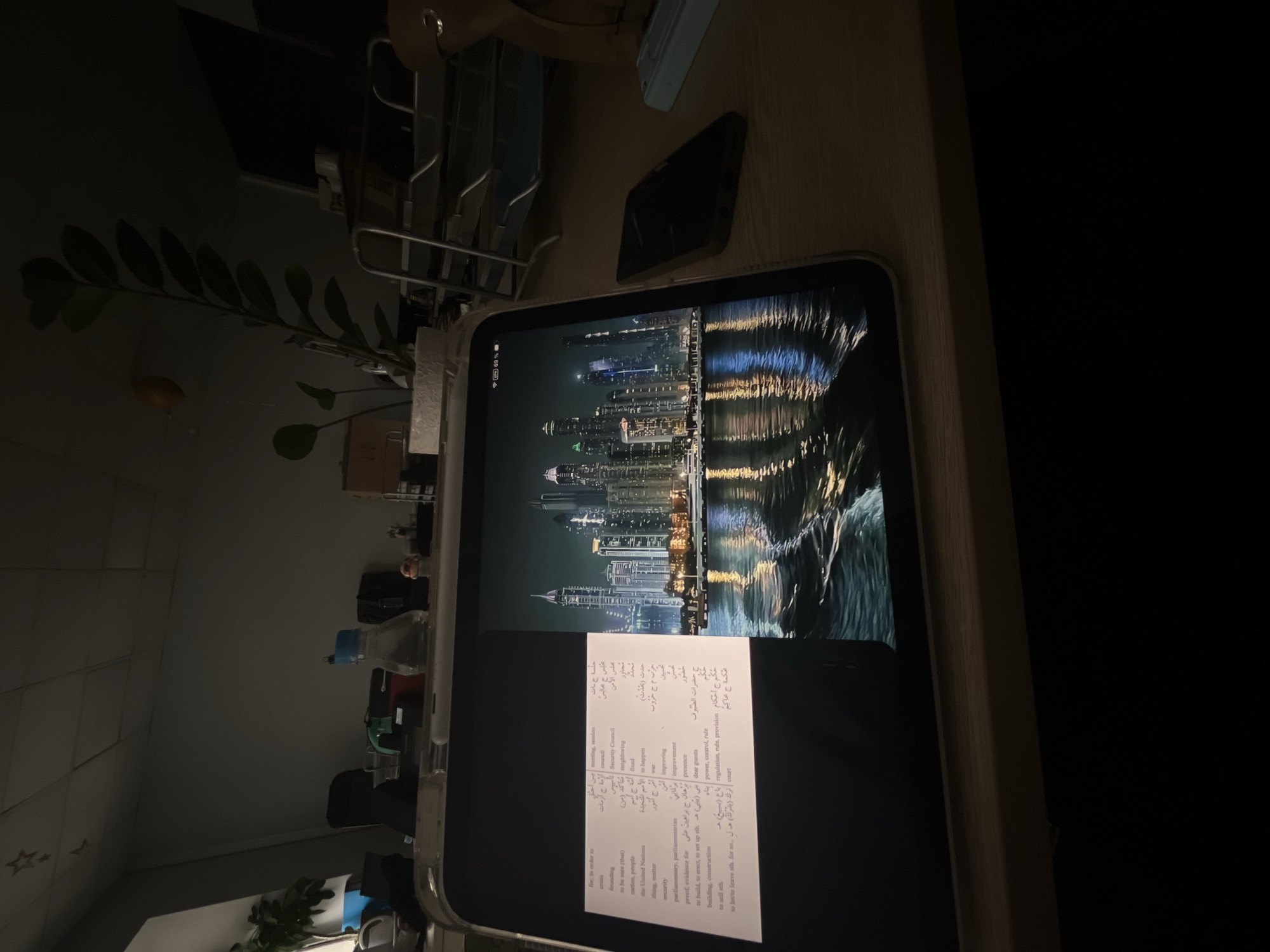 A low-angle, indoor shot depicts a tablet displaying a cityscape at night with a paper overlay containing Arabic text and English translations. The tablet rests on a wooden surface. Behind the tablet, a partially visible storage rack and foliage of a plant are noticeable. The surrounding environment appears dimly lit with the right side of the frame fading into darkness. The image suggests a study or office setting.