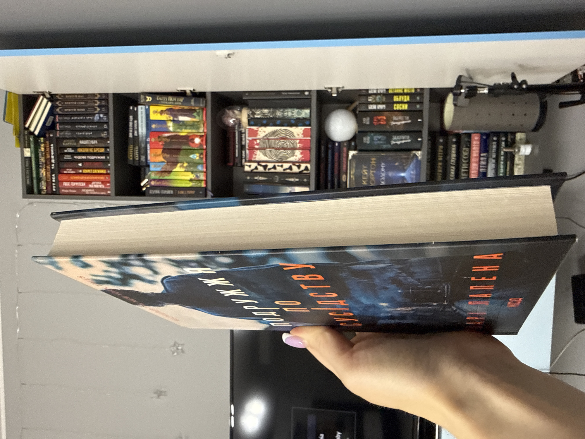 A hand with a manicured nail, painted with white polish and a hint of purple, is holding a thick book with a dark blue cover featuring an orange title. The book appears to be titled "Подружки по сусідству" or a similar Cyrillic script. The background shows a bookshelf filled with many books. One shelf contains colorful spines, while others are filled with books of various colors and sizes. A small, spherical white object is visible on one shelf, and a partially opened cabinet door is on the right side of the image. The environment suggests an indoor setting, possibly a room with a TV or monitor visible in the blurred background to the left. There is no indication of the city of Łódź, Poland.