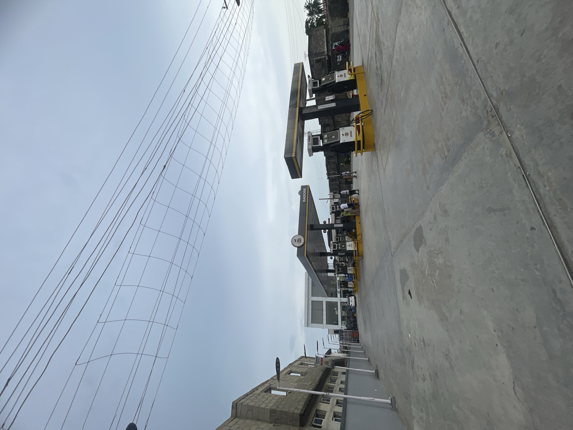 The image displays a gas station under an overcast sky, likely in Ado, Nigeria. A wide concrete forecourt occupies the lower and central portions of the scene. In the mid-ground, two prominent canopies, black with yellow detailing, shelter multiple fuel pumps. The canopy closer to the viewer has the word "BADORE" printed on its side, and a circular sign below it displays "24H," indicating 24-hour operation. The fuel pumps are labeled "SUPER" and "DIESEL," with the "MRS" brand logo also visible on several units.
On the left side of the forecourt, modern streetlights line the concrete path, leading towards a partially constructed building made of concrete blocks. Further back, a contemporary multi-story building with large windows, possibly part of the station's facilities, is visible. In the distance, behind the station, other buildings and palm trees can be seen.
The upper portion of the image shows a vast, light grey sky extensively covered by a dense network of overhead wires, some forming a distinct grid-like pattern. A few people and a white vehicle are present near the fuel pumps, suggesting ongoing activity such as refueling. The scene captures a functional commercial area during daylight hours, characterized by modern infrastructure and clear skies.