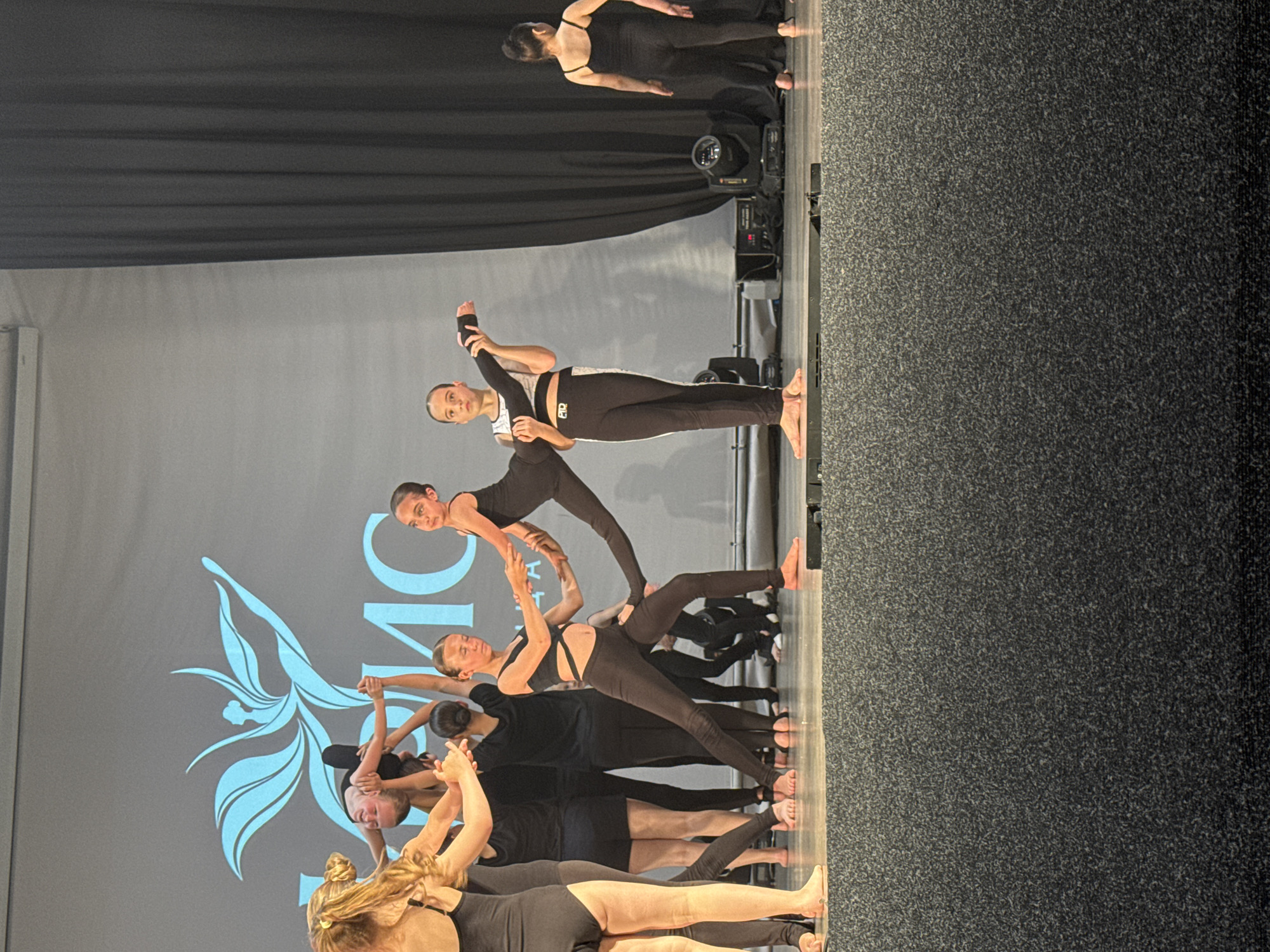 The image depicts a dance performance on a stage, likely at the "Olympic" circus or a similar venue in Sochi, Russia, given the prominent logo resembling the Olympic rings and the lotus flower on the backdrop. A group of young female dancers, dressed in black leotards and leggings, are engaged in a synchronized routine. In the foreground, a dancer is being supported in a lifted pose by two other dancers, her body extended horizontally. To her right, another dancer holds her leg in a graceful pose, her gaze directed forward. Behind them, other dancers are positioned, some in similar black attire, and others appearing to be in transition or preparing for their next move. The stage is illuminated by spotlights, and the backdrop features the aforementioned logo and what appears to be blurred text. The overall atmosphere suggests a performance or rehearsal of a contemporary dance piece.