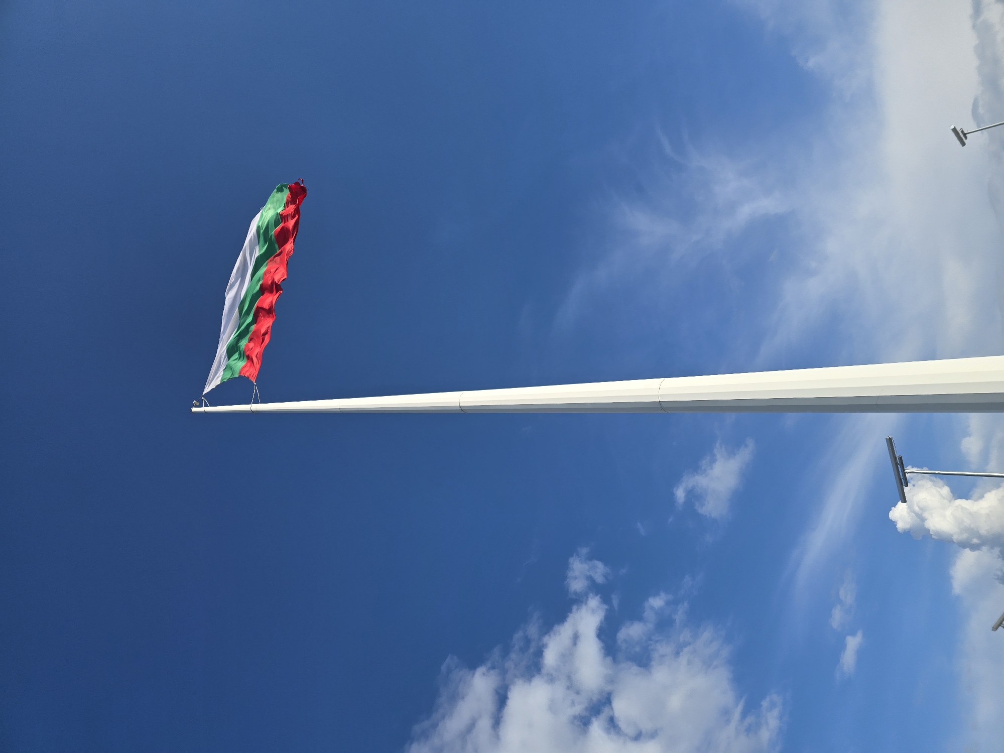 The image shows a Bulgarian flag on a tall flagpole in Yambol, Bulgaria. The flag has three horizontal bands of white, green, and red. The sky is blue, with some white clouds in the background. There are some antenna mounted on the flag pole and clouds near to the antenna.