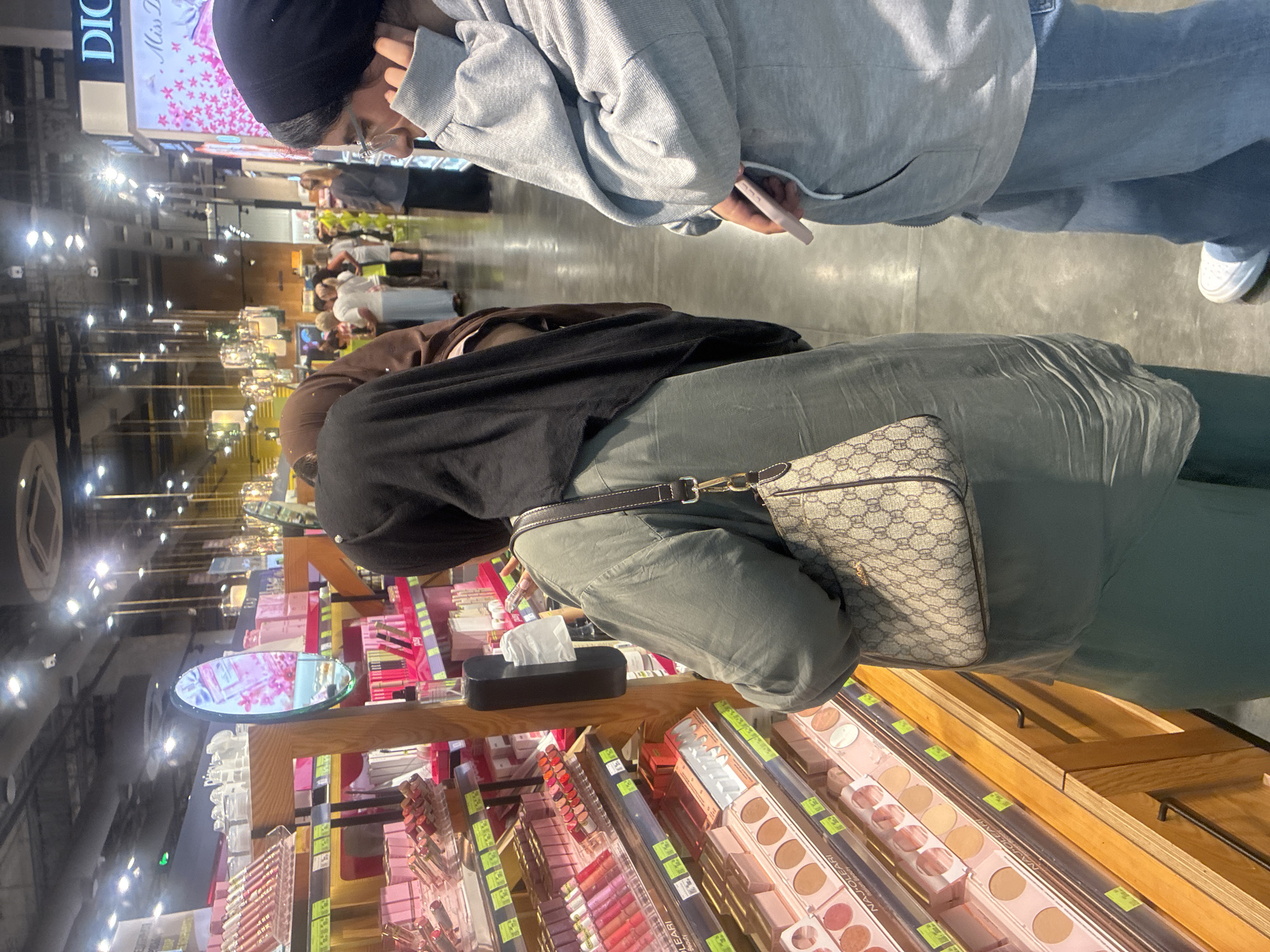In a brightly lit, modern retail store, likely a beauty or department store in Sochi, Russia, several individuals are browsing makeup products. In the foreground, a woman wearing a dark hijab and a green tunic is looking at a display of lipsticks and other cosmetics. She is carrying a patterned handbag. To her right, another person in a grey hooded sweatshirt and blue jeans is holding a pink object, possibly a phone or a compact. Further in the background, other shoppers are visible, some in traditional headscarves, suggesting a diverse clientele. The store features extensive shelving filled with various beauty products, reflecting the consumer culture of a city like Sochi. The overall impression is one of a typical shopping experience in an urban environment, with an emphasis on cosmetics and personal care.