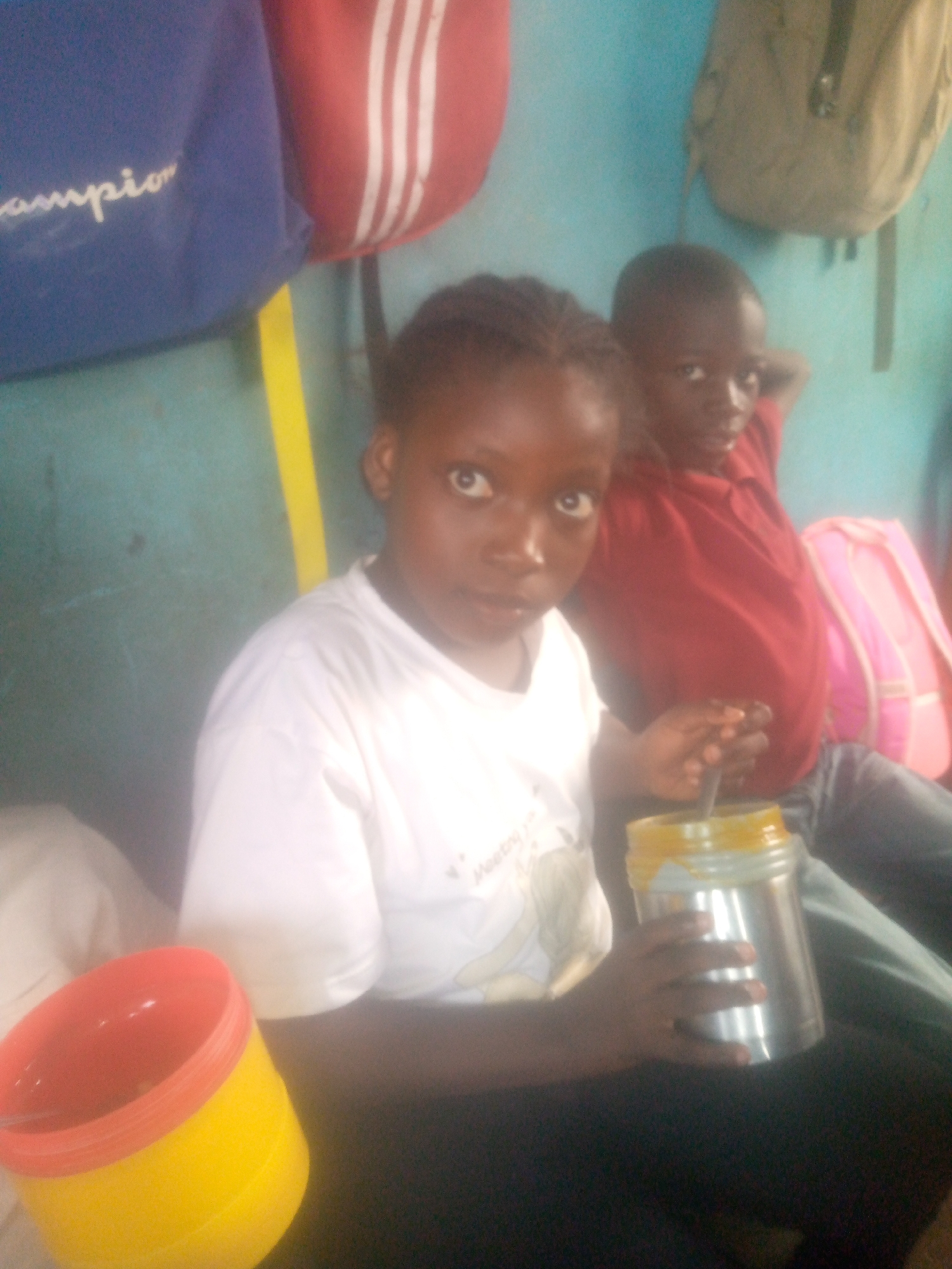 Two children are seated indoors. The child in the foreground is holding a metal container with a yellow-orange substance inside and a yellow and red plastic cup is visible nearby. The second child is sitting behind, wearing a red shirt. School backpacks are hanging on the wall behind them, including one with the word "Champion" printed on it and one that is khaki green. The wall appears to be painted blue with a yellow stripe. The location is Unknown, Unknown.