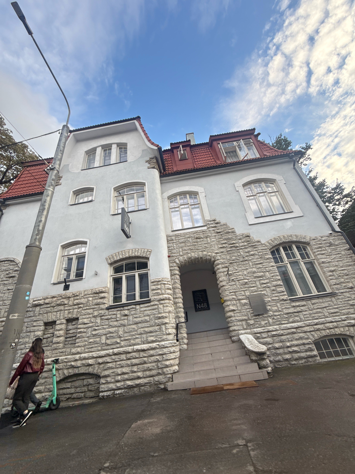 A street-level, low-angle, wide shot captures the exterior of a building with a red-tiled roof, a light blue facade, and a stone base, under a partly cloudy sky. The building appears to be located in Tallinn, Estonia.
In the foreground, to the left, a tall, dark gray lamppost stands at an angle, with a silver metal pole and a dark lamp fixture at the top. Below the lamppost, a woman with long, dark hair, wearing a dark red jacket and dark pants, is walking away from the camera, pushing a bright green electric scooter. She is on an asphalt surface.
The building's facade is a light blue color, with multiple windows of various sizes and shapes. The ground floor and lower sections of the building are covered in a rough, gray stone. A large, arched stone entryway is visible, leading up a set of light gray tiled stairs. A dark sign with white lettering reading "N48" is hanging above the entrance. To the right of the entryway, there are more stone-covered sections and windows.
The background features more of the building's architecture, including dormer windows and a chimney on the red roof. Trees with autumn foliage are visible to the right and behind the building. The sky is a bright blue with scattered white clouds. The overall scene suggests a cool, overcast day in an urban environment.