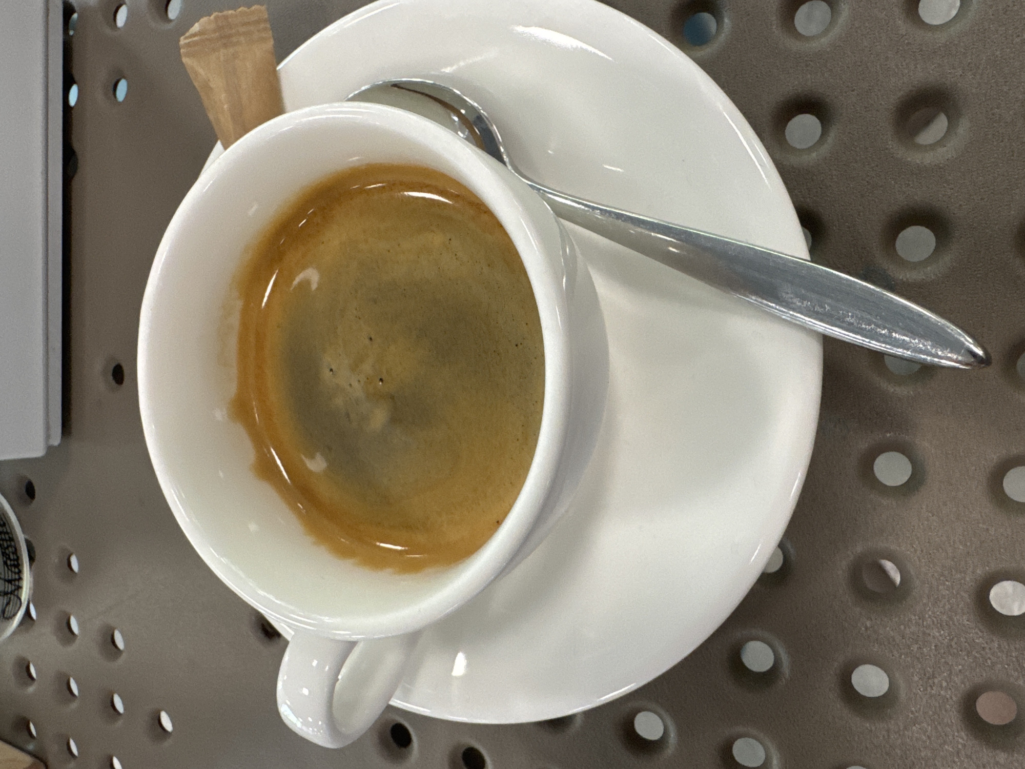 An overhead shot focuses on a white ceramic cup filled with espresso, a saucer, and a spoon on a dark, perforated surface. A small, brown sugar packet is visible to the right of the cup. The espresso has a crema layer. The setting appears to be an outdoor cafe or public space in Orlivshchyna, Ukraine.